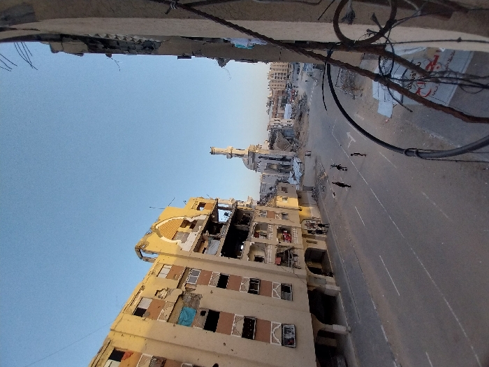 This image depicts a street scene in Khan Yunis, Palestinian Territory, characterized by widespread structural damage under a clear blue sky. In the foreground on the left, a multi-story, light-colored building exhibits significant damage, including blown-out windows, partial wall collapses, exposed interiors, and visible impact marks on its facade. An ornate archway near the top center of this building is also damaged. Directly opposite, on the extreme right, the crumbling facade of another building is visible, revealing exposed rebar and shattered concrete.
A paved road with white lane markings runs through the center of the scene. On the right side of this road in the midground, three individuals, appearing as dark silhouettes, walk away from the viewer. Further down the street, near the base of the damaged building on the left, a small group of approximately three to four people is discernible.
In the background, a heavily damaged mosque with a tall, slender minaret stands amidst further rubble and demolished structures. Beyond the mosque, more distant buildings appear partially destroyed. On the right side of the image, tangled wires and cables hang in the immediate foreground, partially obscuring a white banner with red Arabic text. The text on the banner includes "مستشفى الهلال الأحمر الفلسطيني" (Palestinian Red Crescent Hospital) and "منطقة خان يونس" (Khan Yunis area). The overall environment is marked by debris and the aftermath of significant destruction.