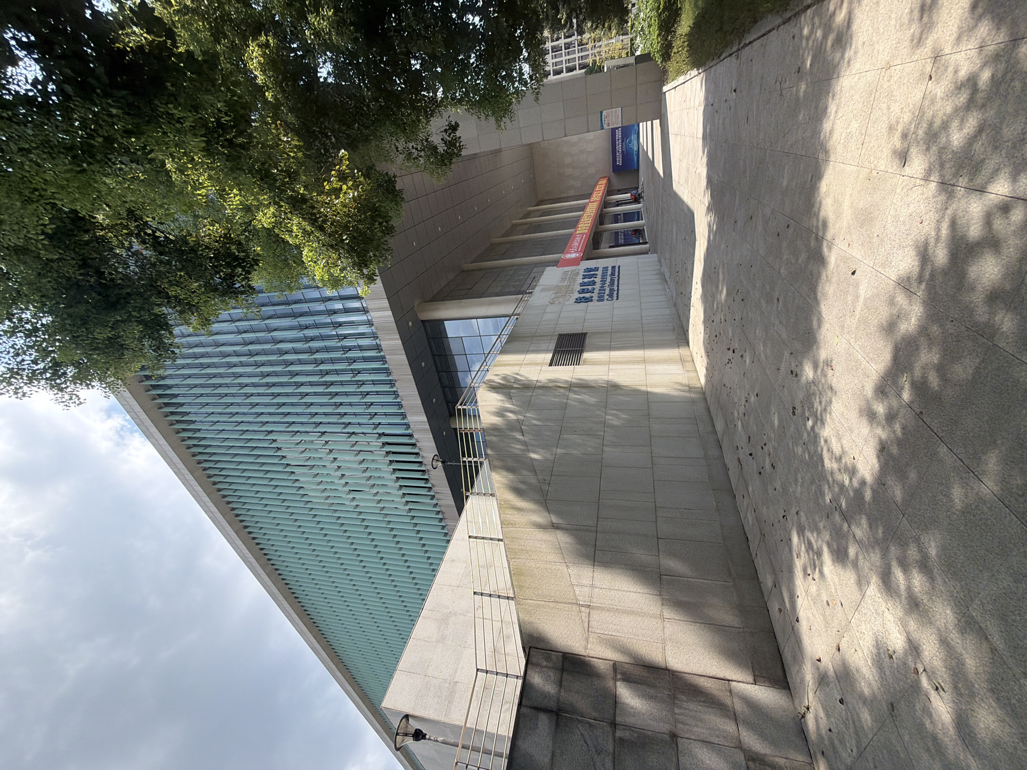 The image shows a modern building with a glass facade, likely a museum or educational institution, in Jiangjin, China. The building has a long, horizontal design with vertical glass panels that create a textured appearance. In front of the building, there is a paved walkway with scattered leaves and dappled shade from trees. To the left, a sloped stone wall leads up to a lower level with a railing and lampposts. A sign with Chinese characters and "College History Museum" is visible on the building's lower level. The sky is partly cloudy. The scene appears to be a daytime view of an outdoor public space.