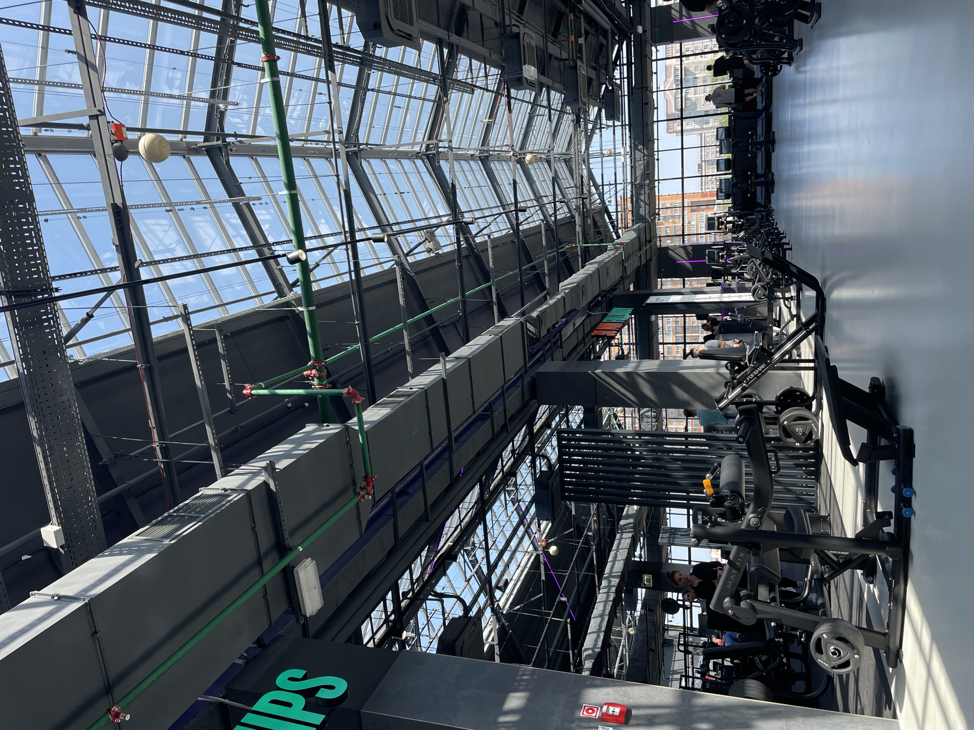 A vertically oriented interior shot depicts a gym setting under a glass ceiling. The ceiling is a grid-like structure of steel beams and glass panes, allowing natural light to filter through. Green pipes run along the underside of the ceiling structure.
On the right side of the image, rows of gym equipment are aligned, with people using the machines. Buildings visible through the windows suggest an urban environment.
There is no information in the image that indicates the location is in the city of Kazan, Russia.
