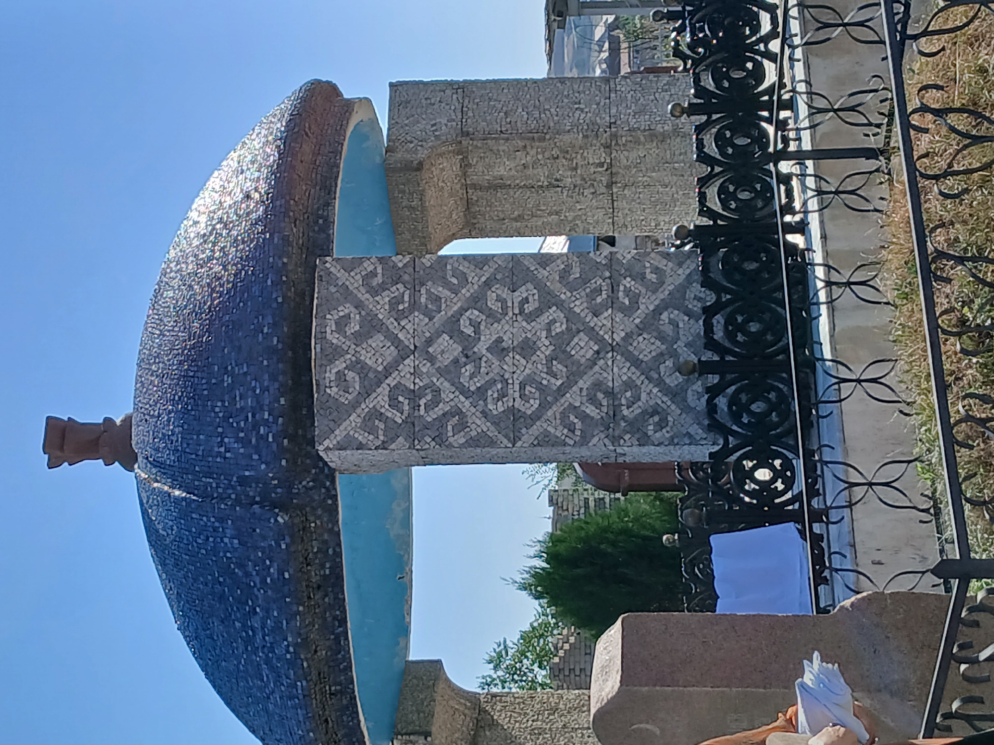 The image shows a large, dark blue tiled dome with a decorative finial on top, supported by stone pillars. The dome is open in the center, revealing a bright blue ceiling. One of the pillars is adorned with a mosaic pattern of interlocking geometric shapes in grey and white. In the foreground, there is a black metal fence surrounding a grave site, with a portion of a pinkish-grey tombstone visible to the left. A white cloth is draped over something behind the fence. In the background, there are trees, brick structures, and a clear blue sky. The scene appears to be a cemetery or memorial site.