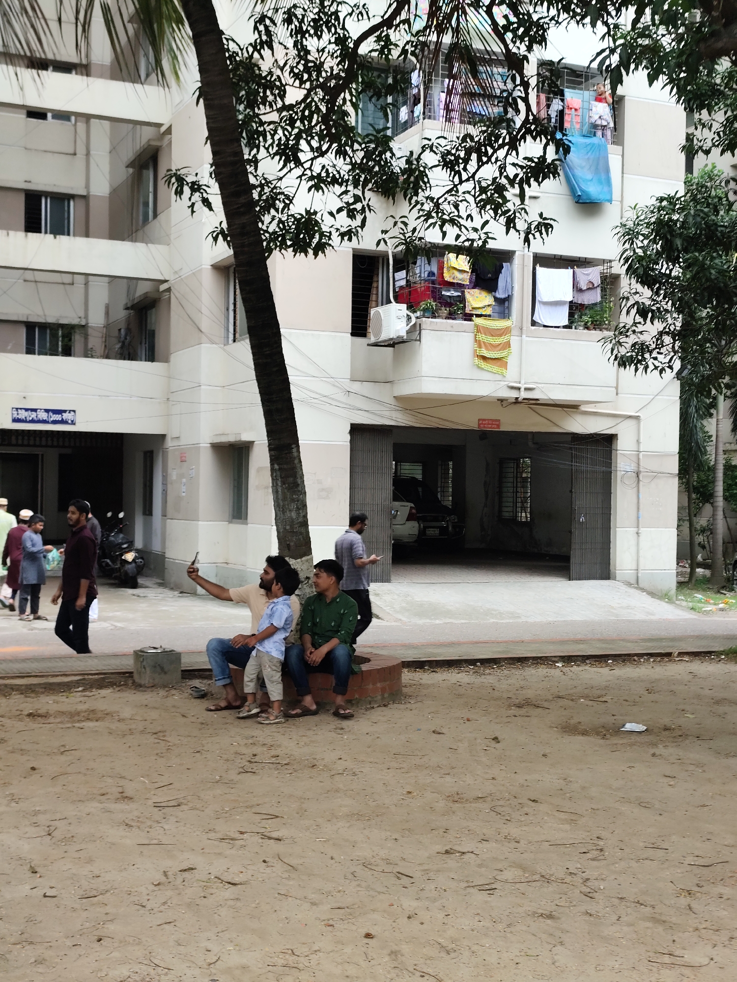 A group of people are seen in front of a building in Dhaka, Bangladesh. The building appears to be an apartment complex with balconies and windows. Laundry is hanging on clotheslines on some of the balconies. In the foreground, three males, one adult and two children, are sitting on a bench. The adult male on the left is taking a selfie with a smartphone. Another adult male is walking in the background. The area in front of the building is a paved pathway and a sandy ground. There are trees and some parked vehicles visible.