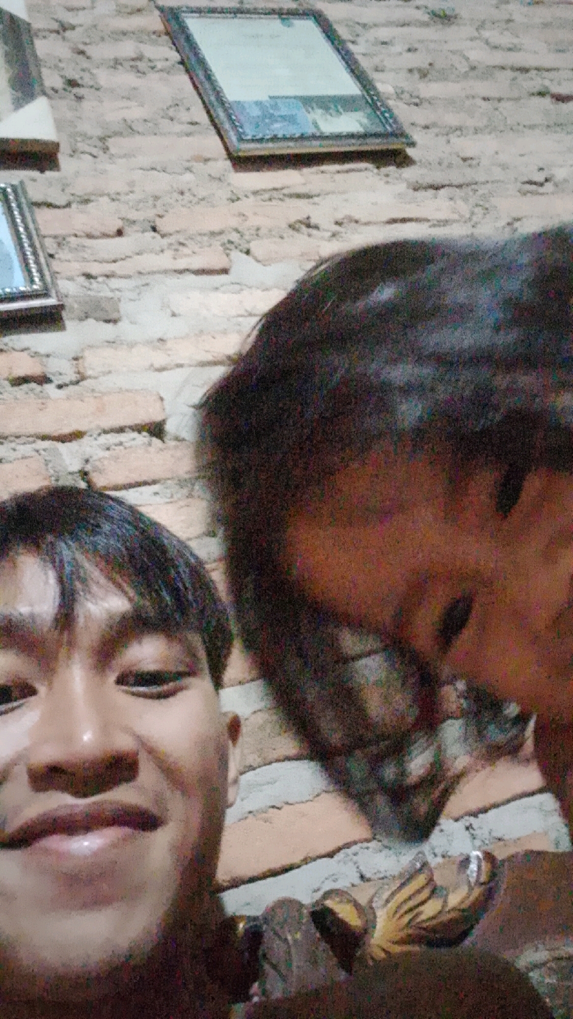In Way Melan, Indonesia, a close-up, slightly blurry photograph captures two individuals in front of a brick wall. The person in the foreground is looking directly at the camera with a smile, their face filling the lower left of the frame. They have short, dark hair and are wearing a dark-colored shirt. To the right and slightly behind this individual, another person's face is visible, partially obscured by darkness and a downward gaze, also with dark hair. Their skin tone appears darker, and they are wearing glasses. The brick wall behind them is rough and textured, with visible mortar lines, and two framed pictures hang on it. One is a landscape orientation, silver-framed picture to the left, and a larger, dark-framed certificate or diploma hangs above and to the right. The lighting is dim, casting shadows and contributing to the image's grainy quality.