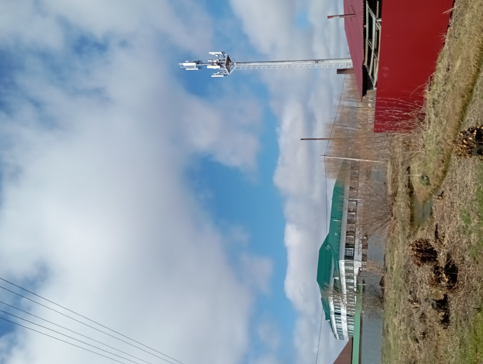 The image shows a scene in what appears to be a residential area. There is a tall telecommunications tower with antennas against a blue and cloudy sky. The tower stands near a red building with a dilapidated appearance. Further in the background are houses with green roofs. A grassy area with some dry vegetation is visible in the foreground and to the side of the red building. Power lines stretch across the lower left portion of the frame.
The location appears to be Yurgamysh, Russia, due to the architectural style of the houses and buildings.