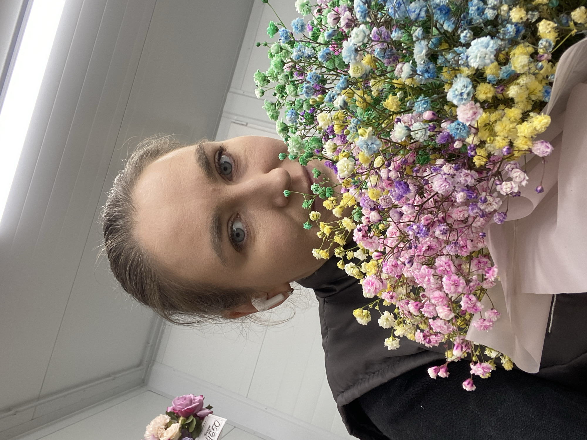 The image shows a person holding a bouquet of colorful flowers in front of their face. The person has light skin, gray hair, and blue eyes, and is wearing a black shirt with a gray vest and a white wireless earbud in their left ear. The bouquet contains flowers in shades of green, blue, yellow, purple, pink, and white. In the background, a white ceiling with fluorescent lights is visible, along with another smaller bouquet of flowers. It is not possible to infer that the photo was taken in Yekaterinburg, Russia.