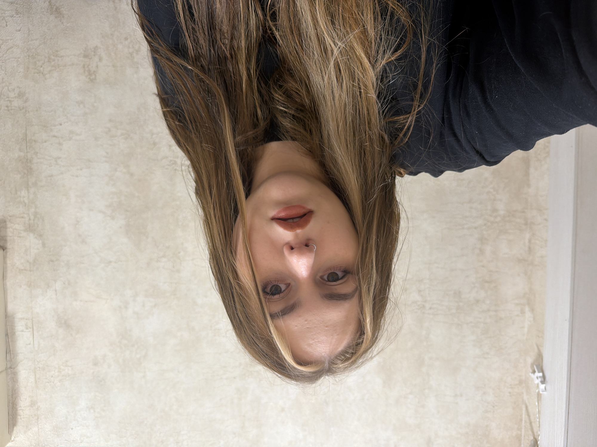 A young woman with long, wavy, brown hair with blonde highlights is looking at the camera. She is wearing a black long-sleeved shirt and has a nose ring. Her makeup includes eyeliner and reddish-brown lipstick. She is positioned in front of a textured, light-colored wall. The environment appears to be indoors, possibly a room. No specific landmarks or indications of Chaikovsky, Russia are visible.