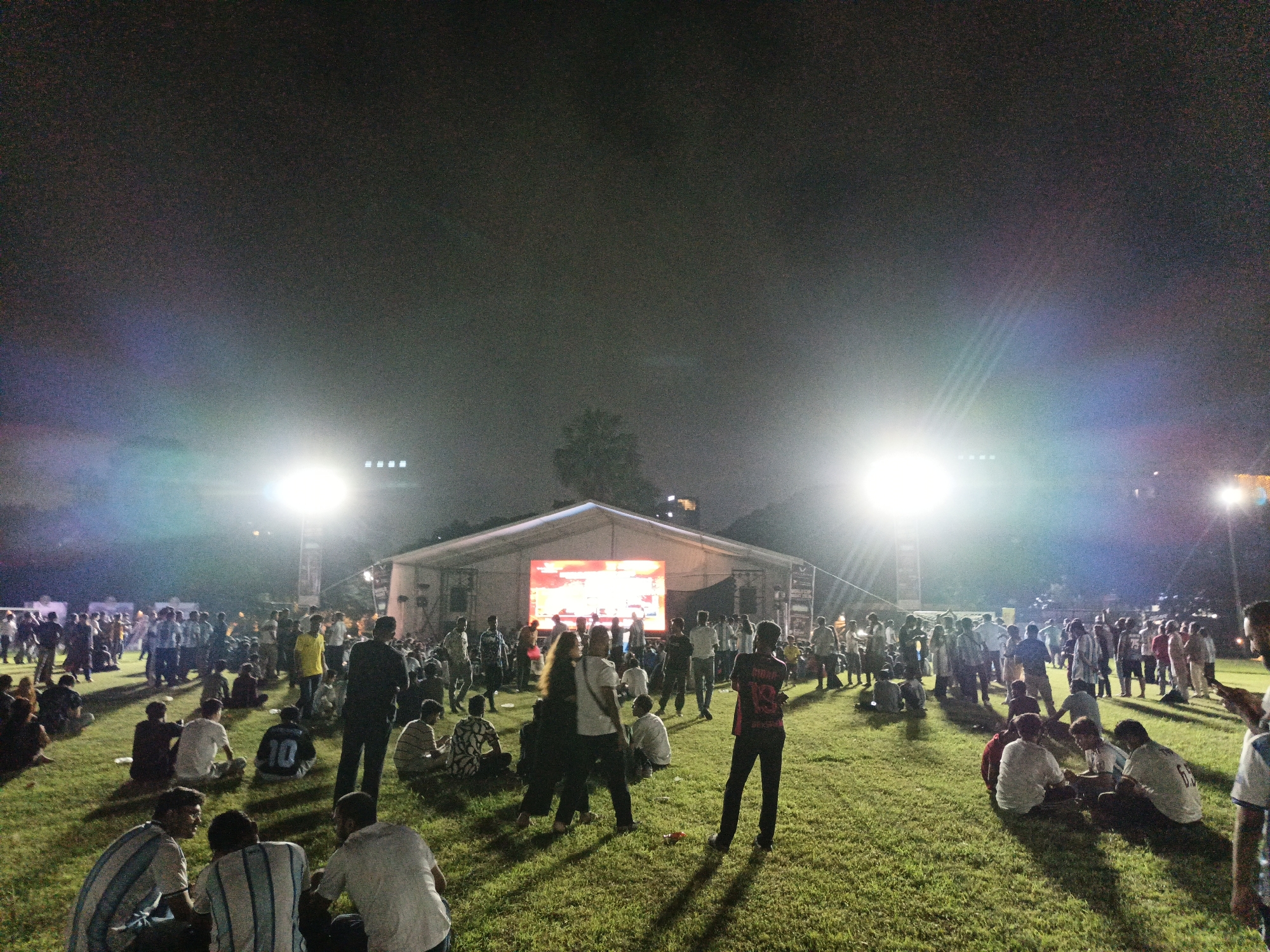 The image captures an outdoor event at night, likely in Dhaka, Bangladesh, given the context. A large, tent-like structure with a bright LED screen displaying an orange graphic serves as the focal point. The screen is illuminated by stage lighting, creating a vibrant atmosphere. Numerous people are scattered across a grassy field, some sitting and others standing. Many are dressed casually, and a few are wearing what appear to be sports jerseys. Bright stage lights are positioned on either side of the tent, casting long shadows and illuminating the crowd. The overall impression is one of a lively gathering or festival.