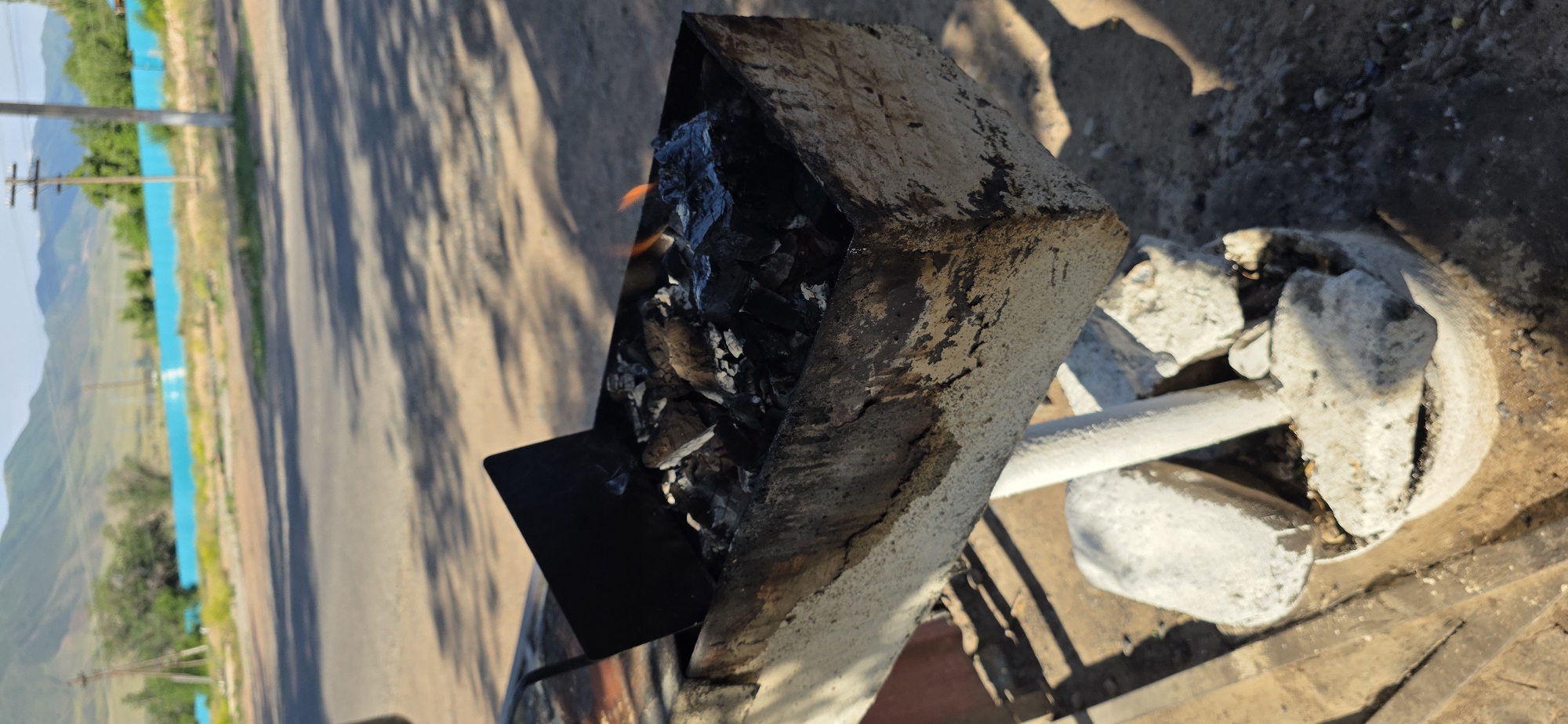 In Sasyk-Bulak, Kyrgyzstan, a close-up shot focuses on a rustic, weathered metal brazier filled with glowing charcoal and small flames. The brazier, positioned outdoors, appears to be set on a concrete base with several large, irregular concrete chunks. A black metal flap is angled within the brazier, partially obscuring the embers. The background shows a paved road with visible tire tracks and shadows cast by an unseen object, suggesting a sunny day. Beyond the road, a vibrant turquoise wall runs along the landscape, backed by dry grass and trees. In the distance, rolling hills form a natural backdrop under a clear sky. A utility pole with wires is also visible. The overall scene depicts an outdoor setting, possibly a roadside or park area, with a focus on the functional, yet aged, cooking equipment.