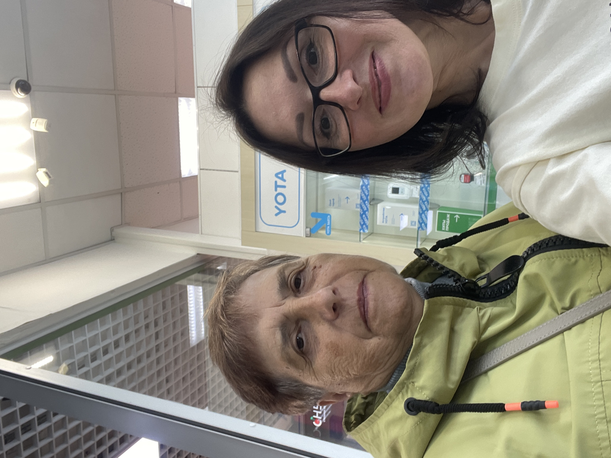 The image is a selfie taken indoors in what appears to be a retail store. Two women are present. The woman on the left has short brown hair, fair skin, and is wearing a light green hooded jacket over a gray turtleneck. She has a gray crossbody bag. The woman on the right has dark brown hair, fair skin, and is wearing glasses. She is wearing a cream-colored t-shirt. Behind them, there is a display shelf with various items and a blue sign with the word "YOTA" in white lettering. The ceiling features fluorescent lighting and what appears to be a security camera. The overall environment suggests a commercial setting, possibly a mobile phone store or electronics retailer, in the city of Kirov, Russia.