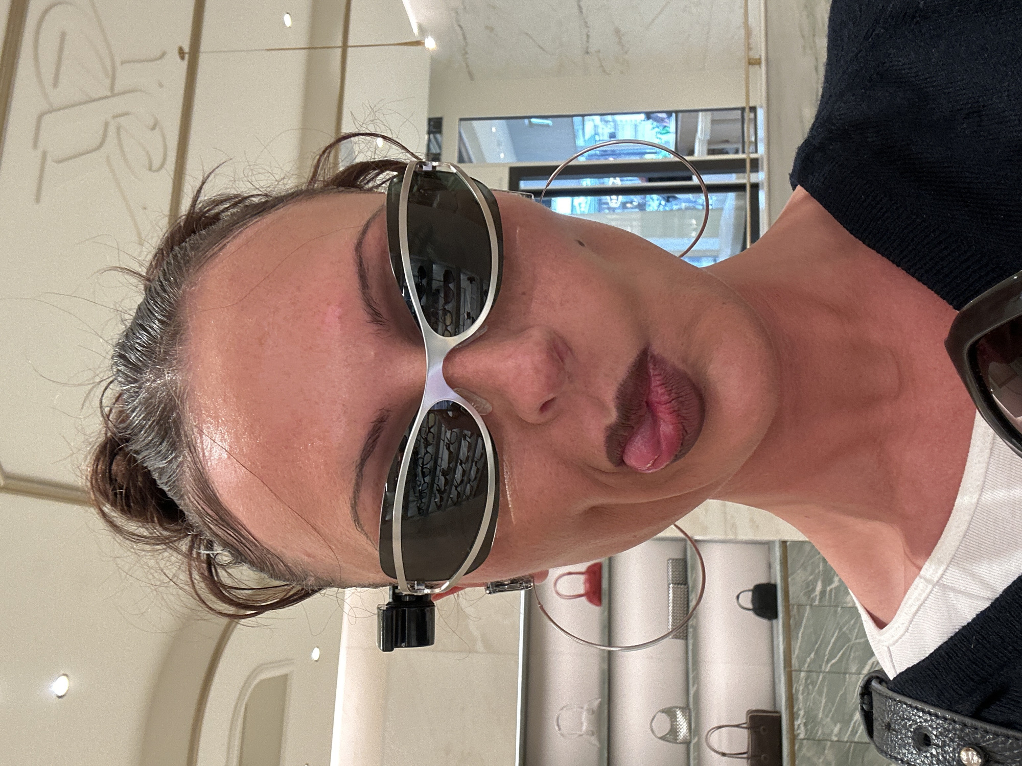 A woman poses in an eyeglasses store, wearing sunglasses. The store interior is visible in the background. The location is likely in London, United Kingdom, but not explicitly confirmed.