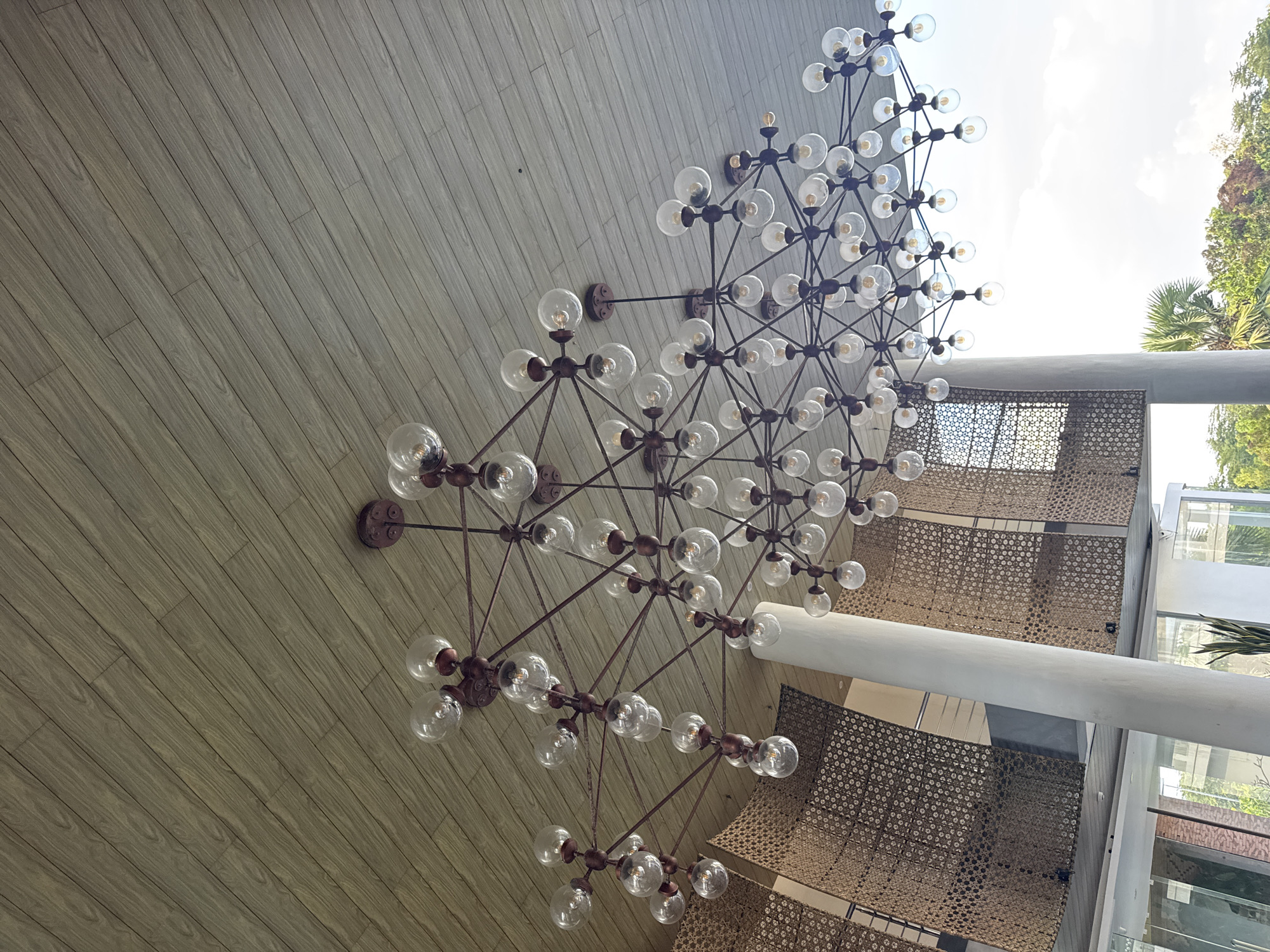 The image depicts the interior of a building in Pecatu, Indonesia. A large, multi-tiered chandelier with numerous clear glass bulbs and a bronze-colored metal frame dominates the foreground. The chandelier is situated against a backdrop of light brown wooden paneling on one side and a more open area on the other. This open area features textured, woven screens, a large white support column, and views of greenery and the sky outside. The image is taken from a low angle, looking upwards towards the chandelier and the surrounding environment.
