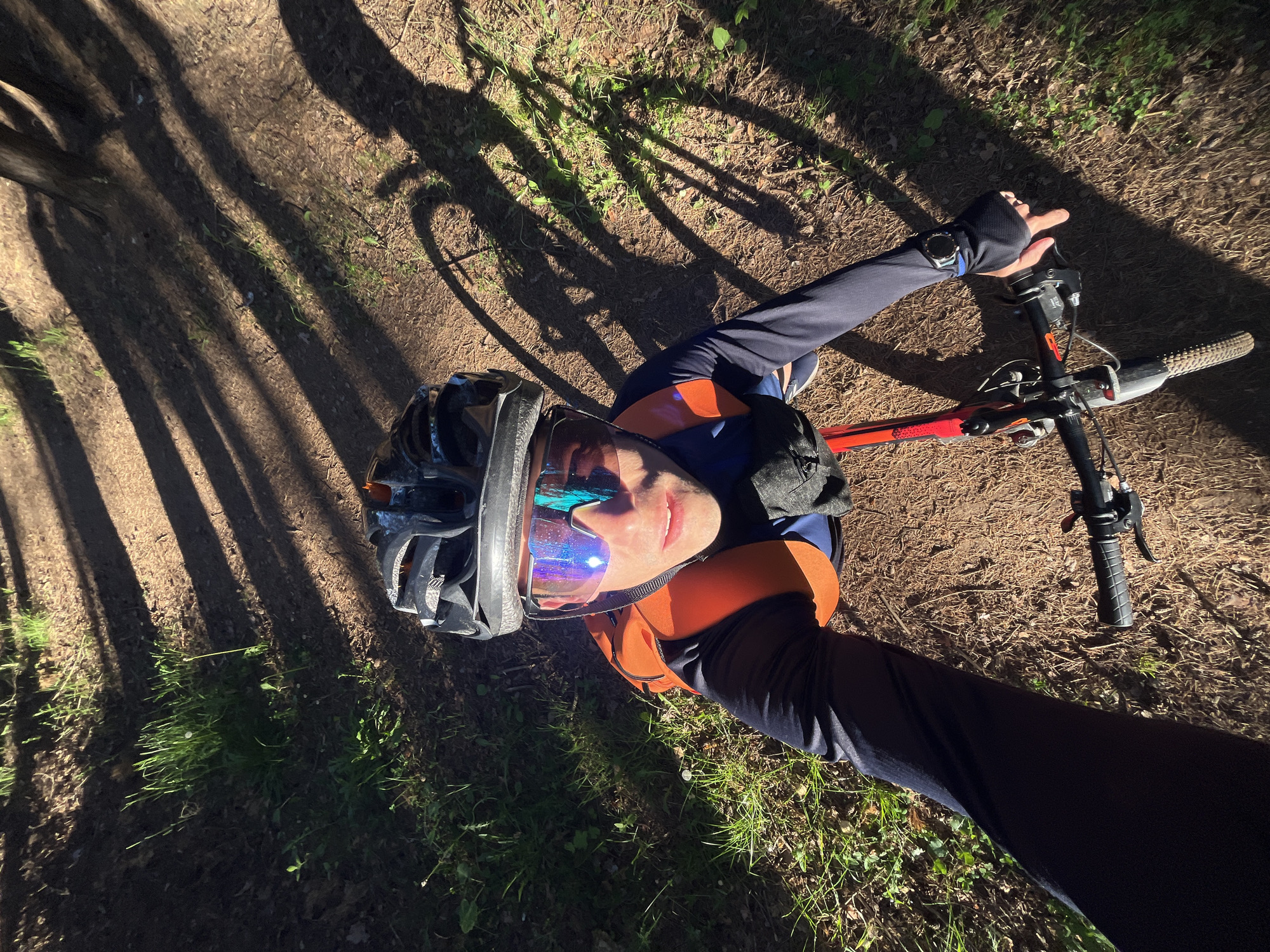 A first-person perspective, point-of-view shot shows a person on a bicycle wearing a helmet and goggles, on a dirt path in a wooded area. The path is dappled with sunlight and shadows from trees. The person is wearing a long-sleeved navy blue shirt with an orange vest over it, and black pants. The bicycle has a red frame and black handlebars. The image appears to have been taken in the city of Chaikovsky, Russia.