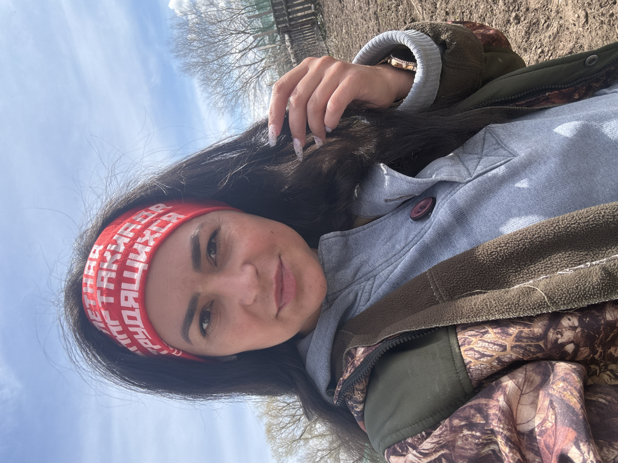 A woman with long, dark hair is looking at the camera. She is wearing a red headband with white lettering, a light gray hooded jacket, and a camouflage jacket. Her nails are long and decorated. The background appears to be an outdoor setting with trees and a blue sky, possibly located in Кемуль, Russia. The environment looks rural or semi-rural.