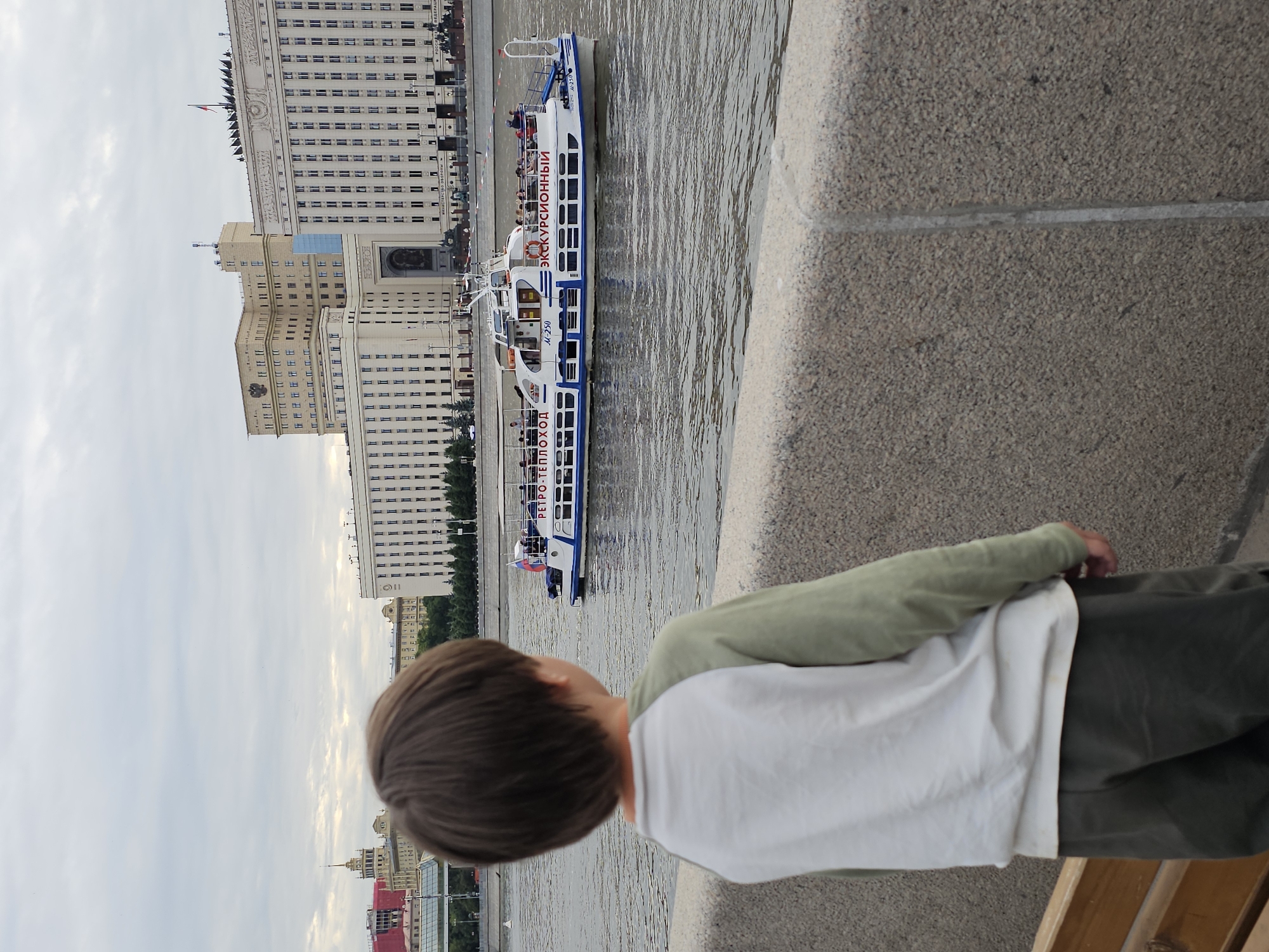 A young boy with dark hair is facing away from the camera, looking towards a wide river. He is wearing a green long-sleeved shirt over a white t-shirt and dark pants. Beside him, on the right, is a stone embankment.
Across the river, a large, multi-story building complex with many windows stretches across the background. The building is light-colored and appears to be of classical architecture. A portion of the building is adorned with decorative elements and flags.
A white tour boat with blue trim is navigating the river. The boat has multiple decks and windows, and people can be seen on board. The word "ЭКСКУРСИОННЫЙ" (Excursion) is prominently displayed on the side of the boat in red lettering, along with the designation "M-250" and "РЕТРО-ТЕПЛОХОД" (Retro-Steamboat).
The sky is overcast, suggesting a cloudy day. The overall scene captures a moment of observation, with the boy seemingly engaged in watching the boat and the cityscape. The presence of the large building and the river suggests an urban setting, consistent with the location context of Moscow, Russia.