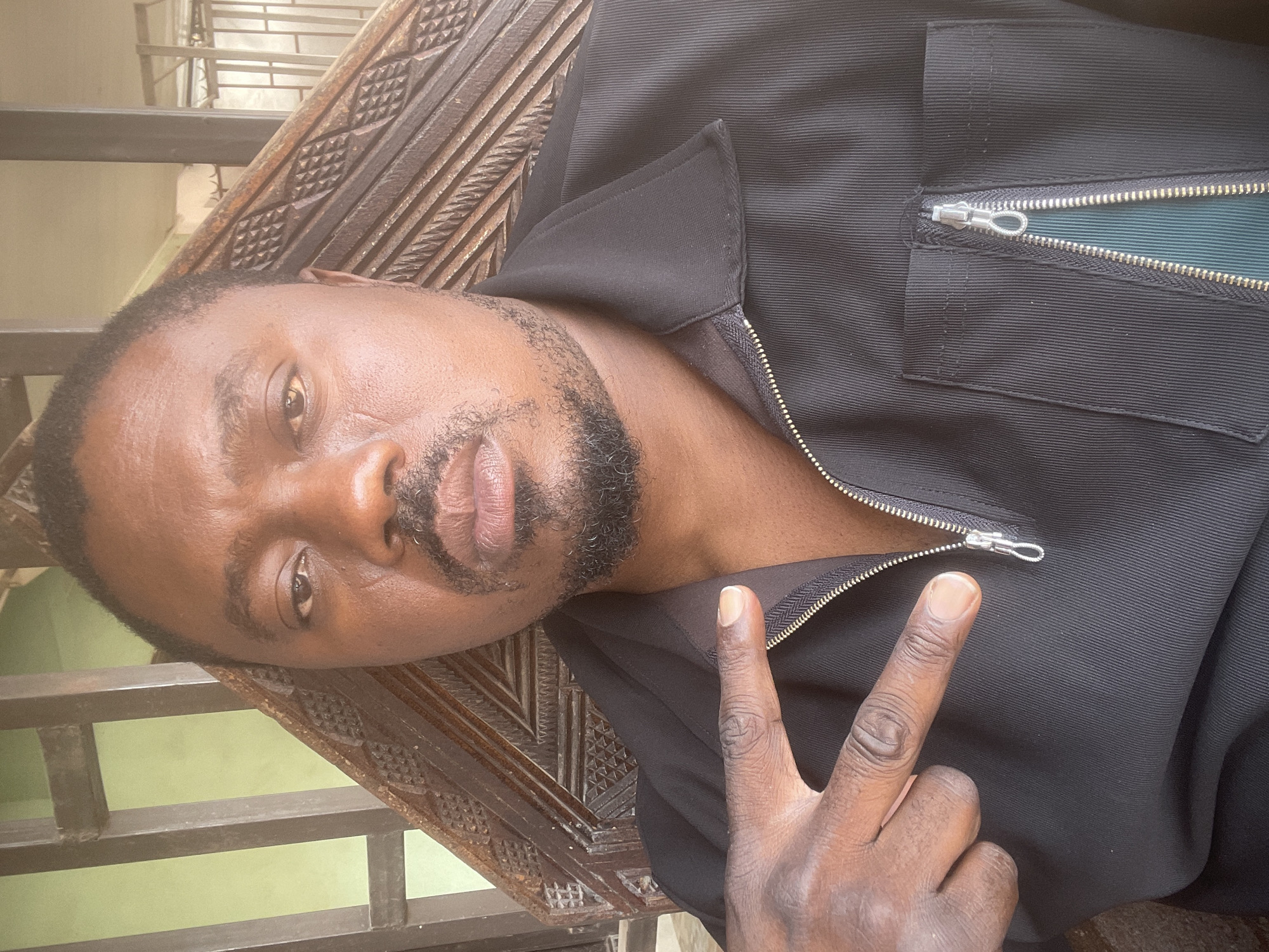 The image depicts a man lying at an angle on a carved wooden structure, likely a bench or platform, with a metal railing visible in the background. The man is wearing a black jacket or shirt with a zipper, and he is making a peace sign with his right hand. He has dark skin, a beard, and short hair. The image is taken indoors, possibly in a home or building in Barakin Akawo, Nigeria, given the prompt's indication. The lighting is fairly even, but the details of the surrounding environment are limited due to the close-up framing of the subject.