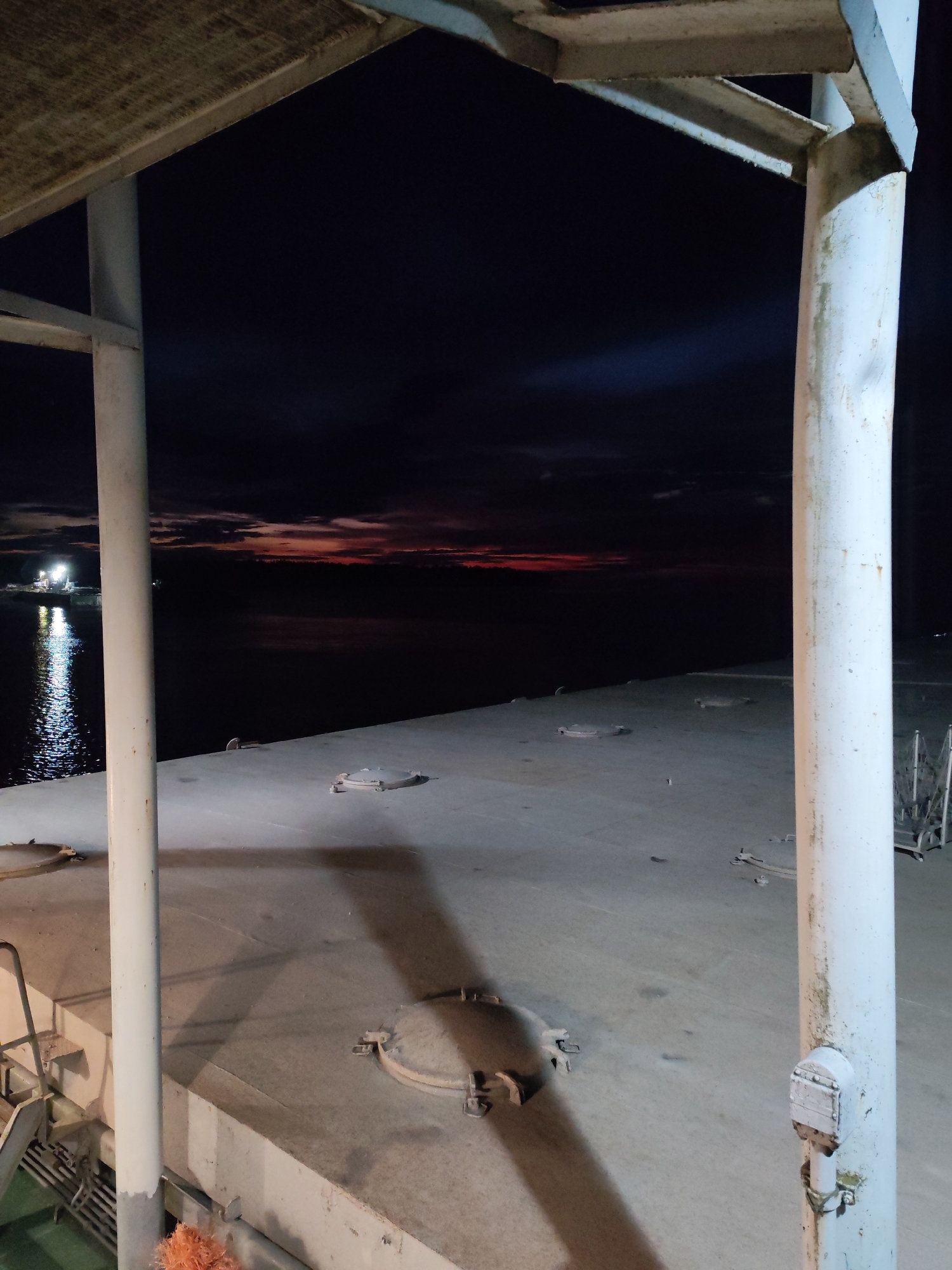 The image depicts an outdoor scene at night on what appears to be a pier or a deck of a vessel. In the foreground, there are two large white cylindrical poles supporting an overhead structure, part of which is visible at the top of the frame. The surface underfoot is a concrete or metal deck with several circular hatches embedded in it. To the left, a metal staircase leads down to a lower level, and a hint of green material is visible below the stairs. In the middle ground, the deck extends towards a dark body of water. On the far side of the water, a distant shoreline is barely visible against the dark sky. The sky is a deep twilight blue, with streaks of red and orange near the horizon, indicating the presence of a sunset or sunrise. The lights of a distant boat or structure are reflected on the water's surface on the left side of the image. The overall atmosphere is dark and quiet, with artificial lighting illuminating the immediate foreground and reflecting on the water. This scene is situated in the city of Kvartsitny, Russia.
