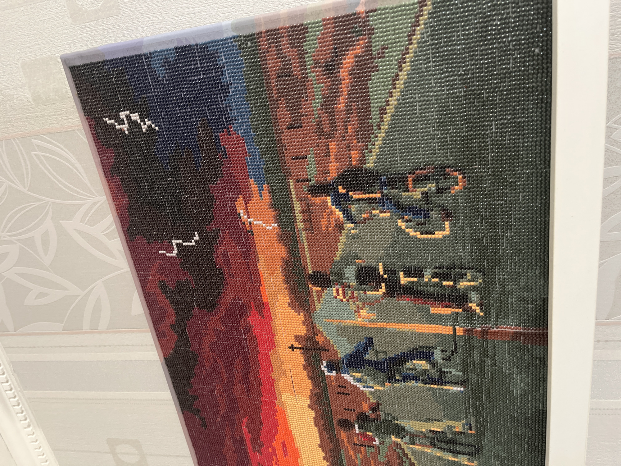A framed diamond painting depicting a cityscape with buildings, water, and boats. The style is blocky and pixelated due to the diamond art format. The background features a gradient of colors, transitioning from dark shades to orange and green. There are some white markings that resemble seagulls. Some of the buildings seem to have text on them. While specific landmarks cannot be identified, based on the boats and the type of buildings, the location is likely Saint Petersburg, Russia.