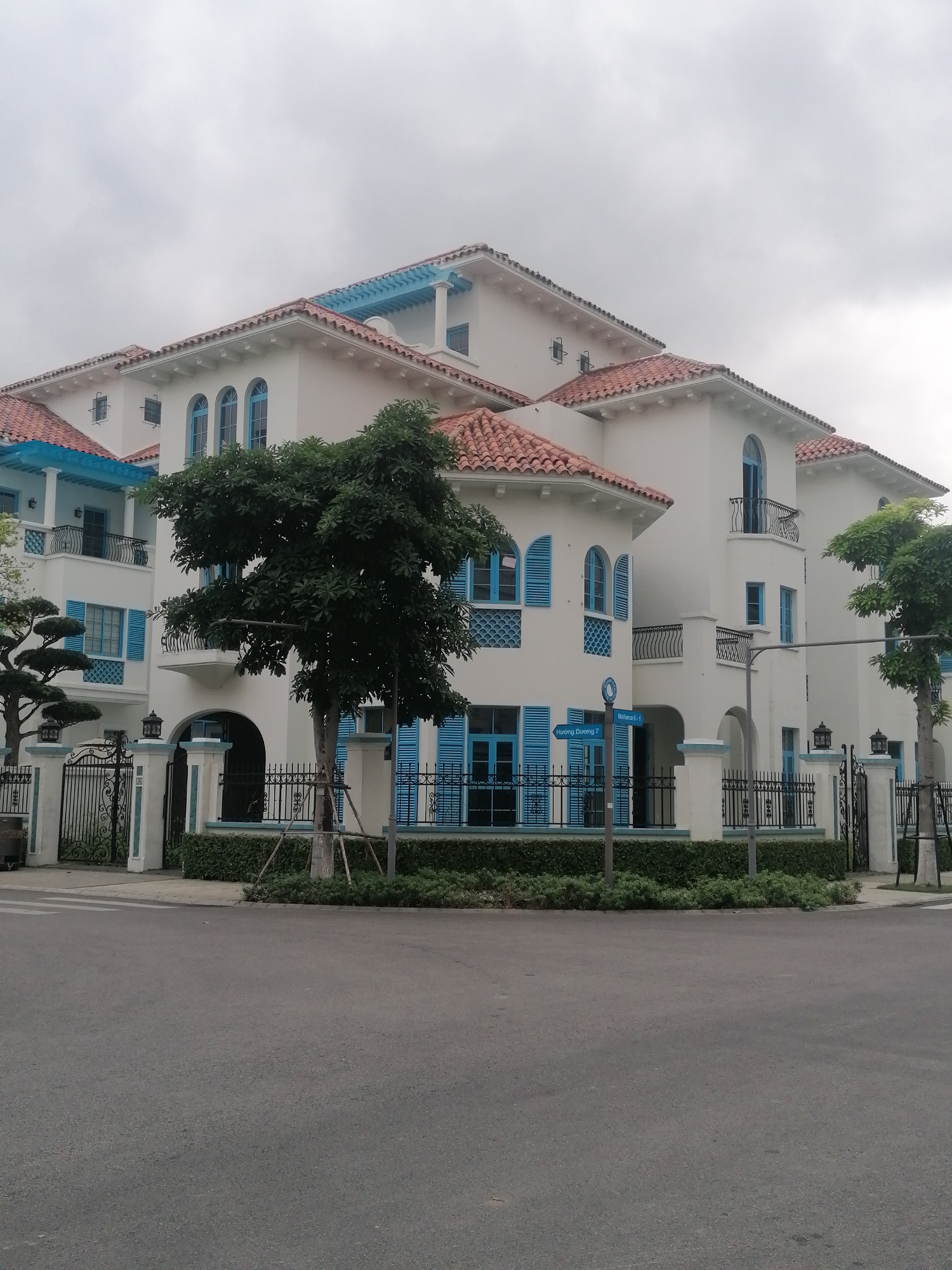 This image presents a street-level view of a large, multi-story building characterized by a distinct Mediterranean architectural style, situated in what appears to be a residential or resort area. The building features crisp white stucco walls, traditional terracotta-colored roof tiles, and vibrant blue accents on its window frames, arched shutters, and wrought iron balcony railings. Several arched windows and doors are visible, some with small balconies. A light blue, possibly decorative, pergola-like structure is also visible on an upper roof section.
The foreground is dominated by a paved asphalt road, with a crosswalk partially visible on the right, indicating a street or intersection. The property is enclosed by a black wrought iron fence with decorative posts and matching gates, adorned with light blue trim and lamps. Green hedges are neatly maintained along the base of the fence. Several trees enhance the landscaping, including a prominent, leafy tree in the center foreground supported by wooden stakes, and other smaller, sculpted trees.
The weather appears overcast, with a uniformly gray sky, suggesting a cloudy day or a time with diffused light. No people are present, and no specific activity is occurring, lending a quiet atmosphere to the scene. A blue street signpost displays text in white, reading "Mallorca 6-1" and "Huong Duong 7," likely indicating street names or section numbers within the Bai Chay Ward, Vietnam location.