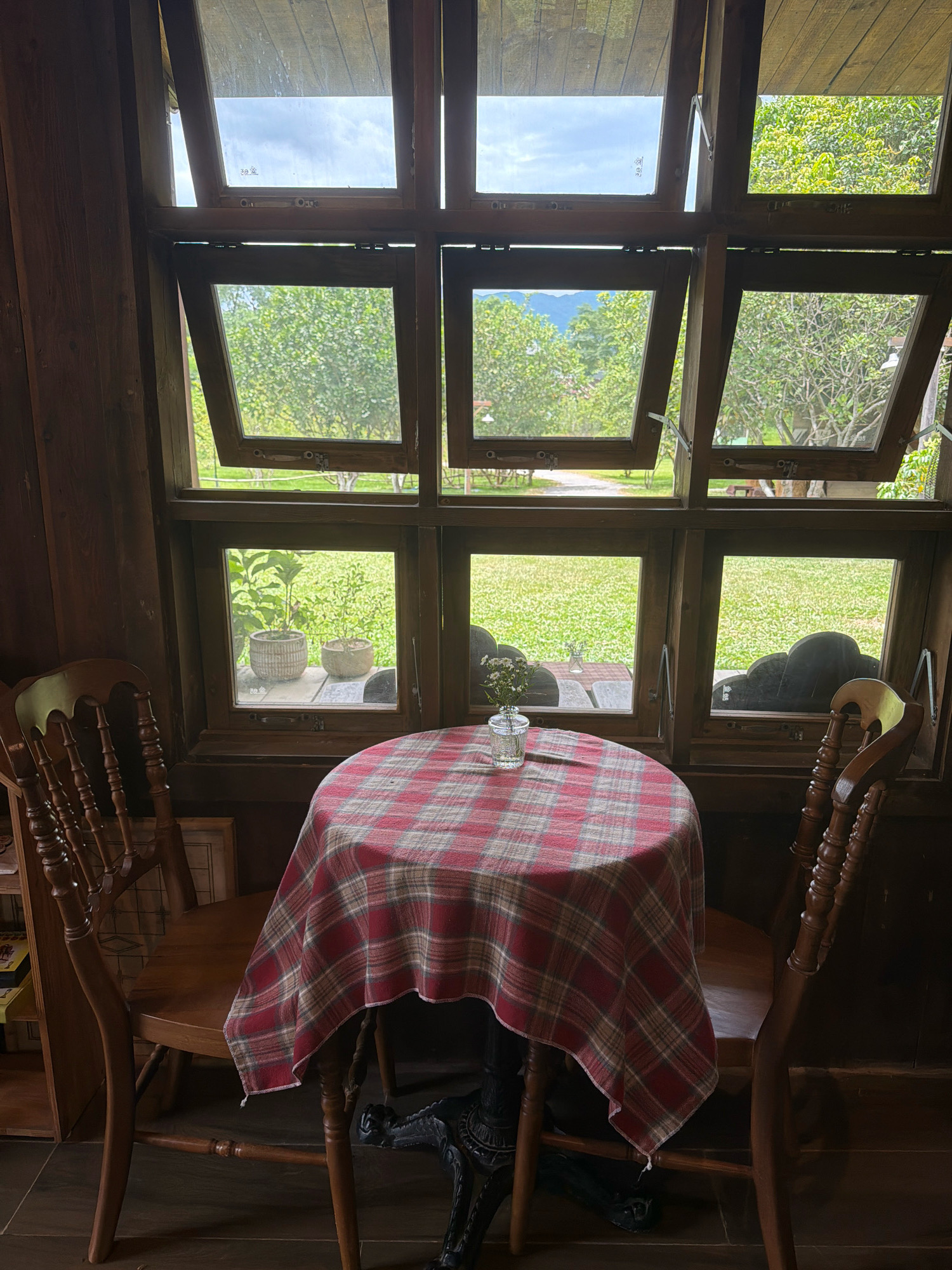 The image depicts an indoor seating area with a view of a lush green landscape through a large wooden-framed window. The window is divided into multiple panes, some of which are open, allowing fresh air and natural light to enter. In the foreground, a small round table is set with a red and plaid tablecloth. A clear glass vase containing a small bouquet of white flowers sits in the center of the table. Two wooden chairs with intricate carvings are placed on either side of the table. The interior walls are paneled with dark wood, creating a cozy and rustic atmosphere. The scene appears to be a quiet corner of a cafe or restaurant, possibly in a suburban or rural setting outside of Đà Nẵng, Vietnam, given the extensive greenery and mountainous backdrop visible through the windows. No people are present in the image.