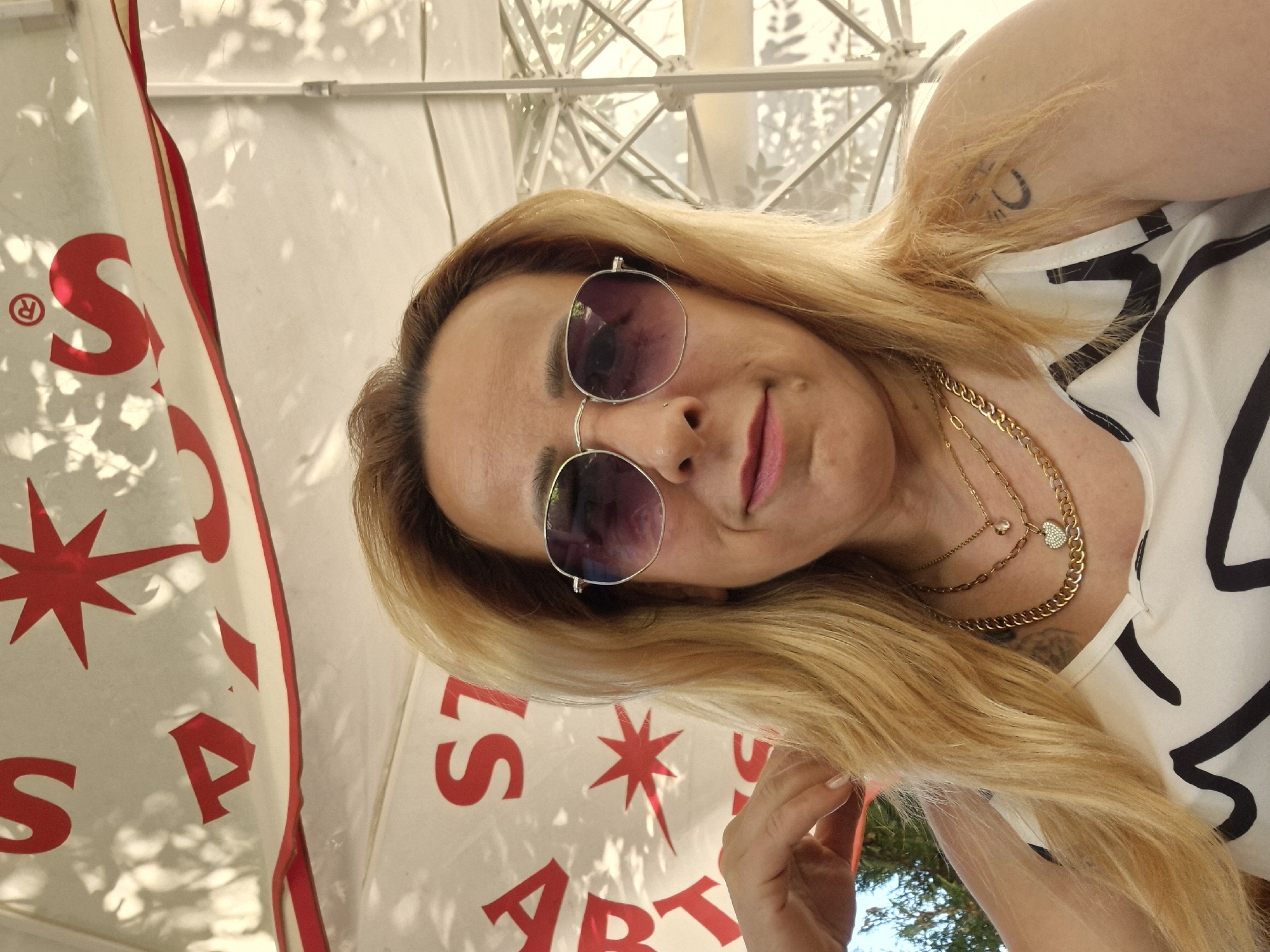 This is a close-up, tilted selfie of a woman with blonde hair and sunglasses. She is wearing a white top with black abstract designs and multiple gold necklaces, one featuring a small heart charm. A tattoo is visible on her upper arm. The background is blurry but appears to be outdoors, with dappled sunlight casting shadows on a white surface, possibly an umbrella or canopy, and glimpses of green foliage and blue sky.
To the left of the woman, a white banner or flag with red lettering and designs is partially visible. The letter "S" with a registered trademark symbol is prominent, along with a red star-like shape. Further down, the letters "S", "P", and "A" are visible. On the right side of the frame, more red lettering is seen, including what appears to be "L S" and the letters "A R T". The overall impression is of a casual, sunny outdoor setting. The woman's expression is relaxed, with a slight smile. The time of day appears to be daytime, judging by the bright sunlight.