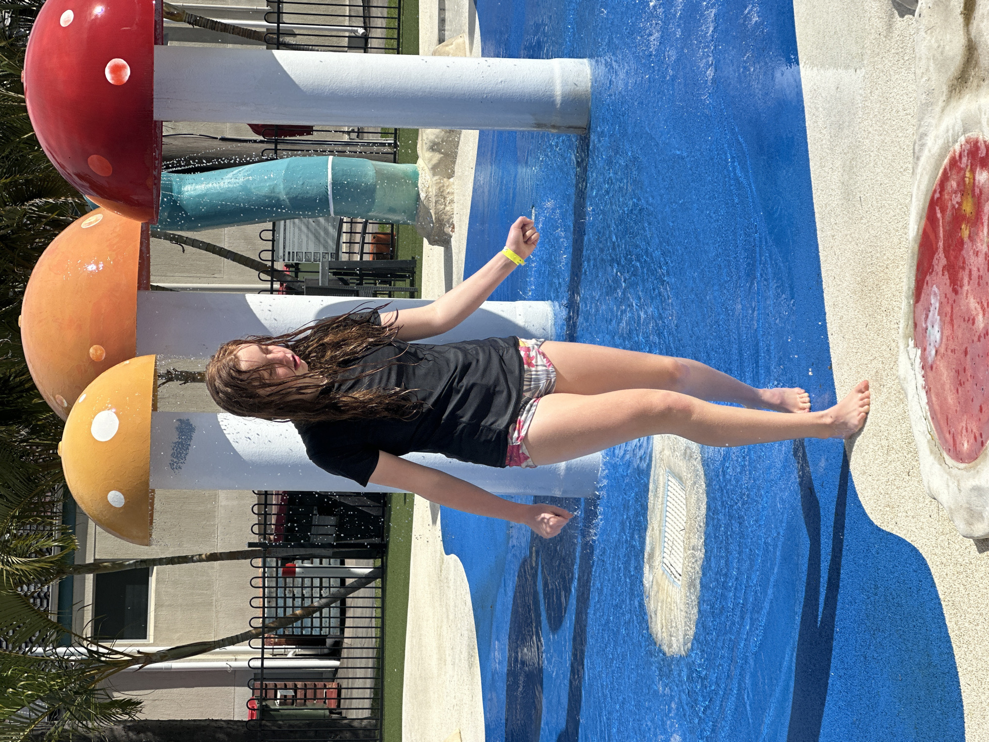 A young girl with wet, shoulder-length brown hair and a black t-shirt stands in a blue water play area, partially obscured by a white pole. She is wearing plaid shorts. The water is shallow, with splashes around her feet and ankles. Behind her, a blue and white patterned cylindrical structure is visible, along with a red mushroom-shaped water feature with white dots. Further in the background, there are more mushroom-shaped water features, including an orange one. Palm trees and a building with a dark window are also present. A black metal fence runs along the edge of the water play area, separating it from a grassy area. The scene appears to be outdoors on a sunny day. The location is the Nambucca Heads Splash Park in Nambucca Heads, Australia.