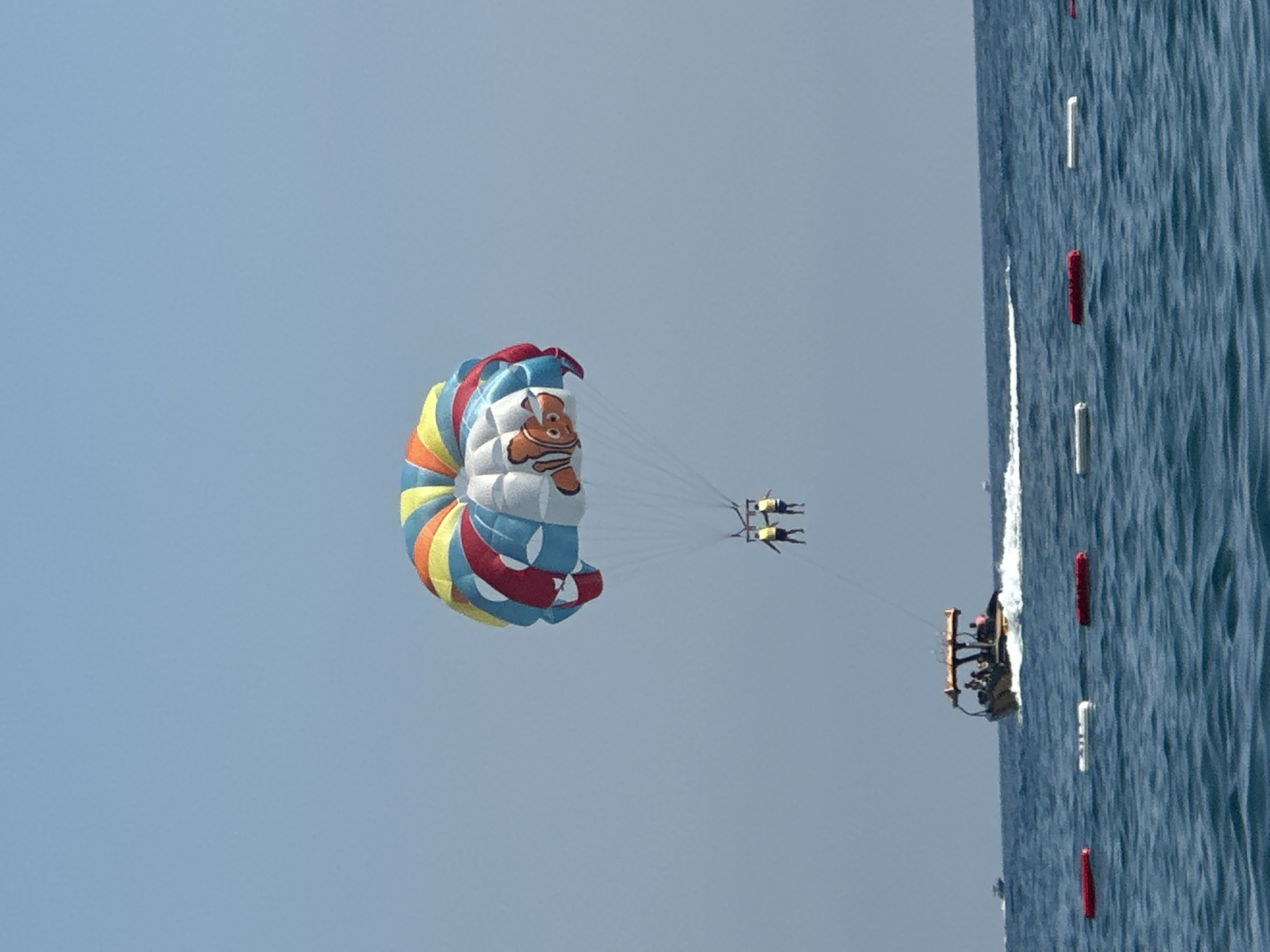 In the city of Boreti, Montenegro, a parasail adorned with a depiction of the character Nemo, is soaring through the bright blue sky. Two individuals, wearing yellow life vests, are suspended beneath the parasail, experiencing the thrill of parasailing. Below them, a motorboat navigates the turquoise waters, creating a white wake as it moves. The sea is dotted with floating buoys, some red and some white, marking a section of the water. The scene is set on a sunny day, with the vast expanse of the sky meeting the undulating surface of the sea.