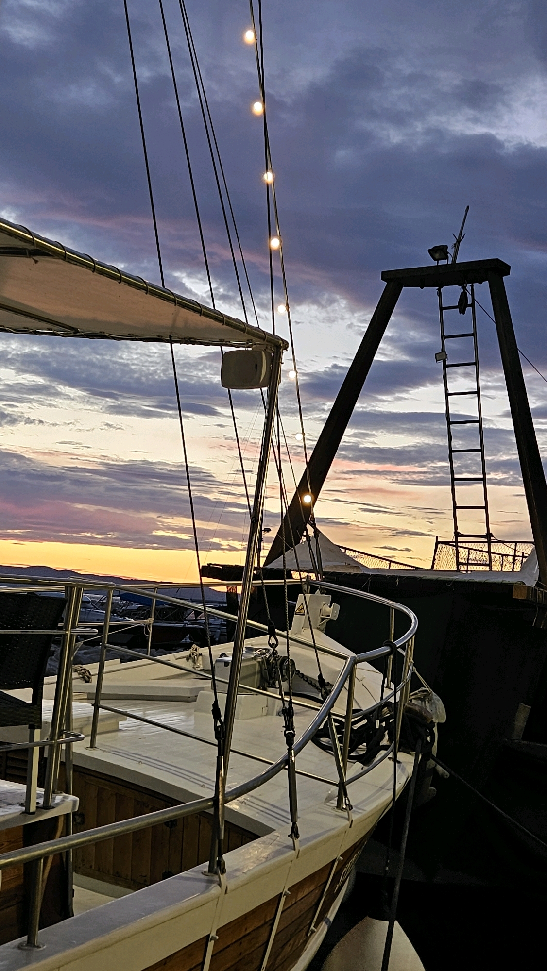 The image shows a close-up of a sailboat docked at dusk, with a dramatic sky filled with clouds in shades of purple, pink, and orange. String lights hang from the rigging, illuminated against the darkening sky. The foreground features the deck of the sailboat, with its white surface, wooden hull accents, and polished metal railings. In the background, a larger, darker vessel is visible, with a ladder leading up to its deck. The setting appears to be a harbor or marina, likely in a coastal town. While the specific location within Općina Tučepi, Croatia is not explicitly shown, the scene suggests a typical Mediterranean harbor setting. There are no people visible in the image.