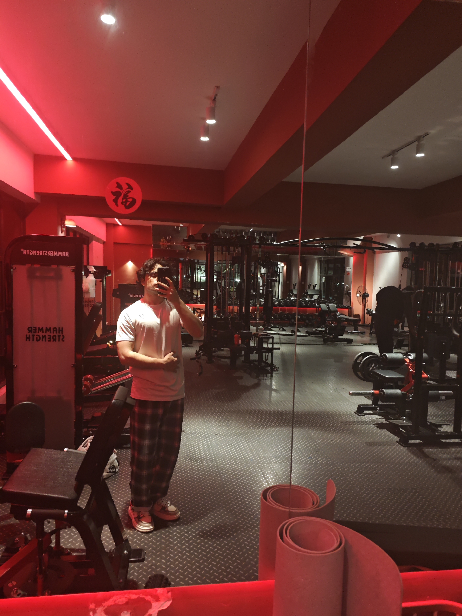 A man stands in a gym located in Chongqing High-tech Zone, China, taking a selfie in front of a mirrored wall. The gym features Hammer Strength equipment and various weightlifting machines. The lighting is predominantly red. The man is wearing a white t-shirt and plaid pants. A rolled-up mat is positioned near the mirror's base.