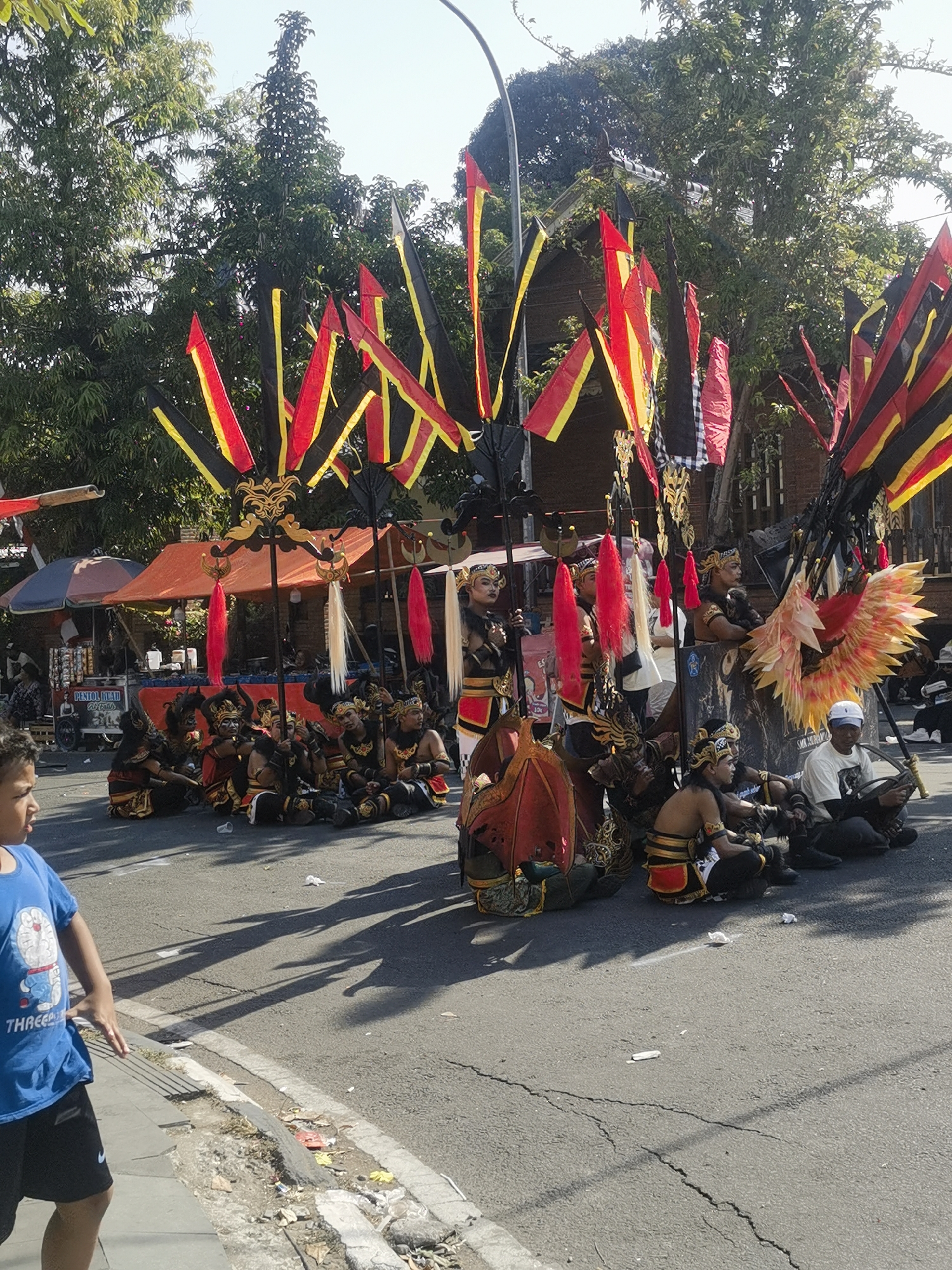 The image depicts a street scene in Blora, Indonesia, likely during a cultural event or parade. In the foreground, a young boy wearing a blue t-shirt with a cartoon character and black shorts stands on a sidewalk, his body turned to the left, suggesting he is looking towards something or someone off-camera.
Behind him, the street is filled with performers in traditional attire, seated on the asphalt. They are adorned in elaborate costumes, featuring headdresses, golden ornaments, and red and black fabrics. Some performers hold tall, decorative poles with red and yellow banners attached, creating a festive atmosphere. Several large, colorful props, including what appears to be a mythical bird or phoenix with fiery wings, are also present.
In the background, tents or stalls are set up along the side of the street, indicating a market or food vendors. Trees line the street, providing shade, and the sky is clear and bright, suggesting a sunny day. The presence of people sitting on the street and the decorated poles suggests a procession or performance is either underway or about to begin. The overall scene conveys a sense of cultural celebration and community gathering in an urban environment.