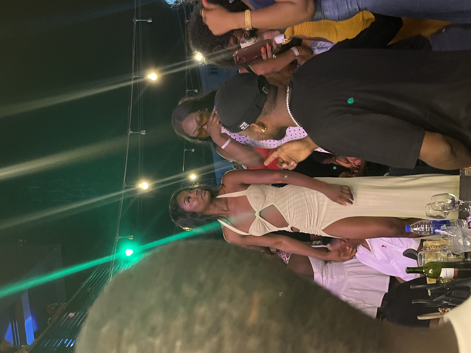 In the dimly lit environment of a nighttime outdoor venue, possibly a party or event, several individuals are captured in a candid moment. The foreground is partially obscured by the out-of-focus head and shoulder of a person. To the left, a woman in a light-colored, form-fitting dress with a thigh-high slit and a cutout detail at the torso stands prominently. She is looking to her right, with her hair dark and styled.
In the midground, a man wearing a black t-shirt, a black beanie, and a diamond necklace is partially visible from the back. He is facing left, with his right hand raised as if gesturing or speaking. Beside him, a woman in a white dress with black polka dots and wearing glasses is also facing left. Further to the right, another woman with dark, curly hair is holding a phone and wearing a gold bracelet. She appears to be interacting with others.
In the background, there are other people, some partially visible, and decorative lights, including a prominent green light beam that cuts across the scene from the upper left. The environment appears to be a social gathering with elements of a stage or elevated area visible in the upper left. On a table in the lower part of the image, there are bottles of wine and water, along with glasses, suggesting refreshments are present. The overall scene depicts a social gathering with a focus on individuals interacting within a festive atmosphere.