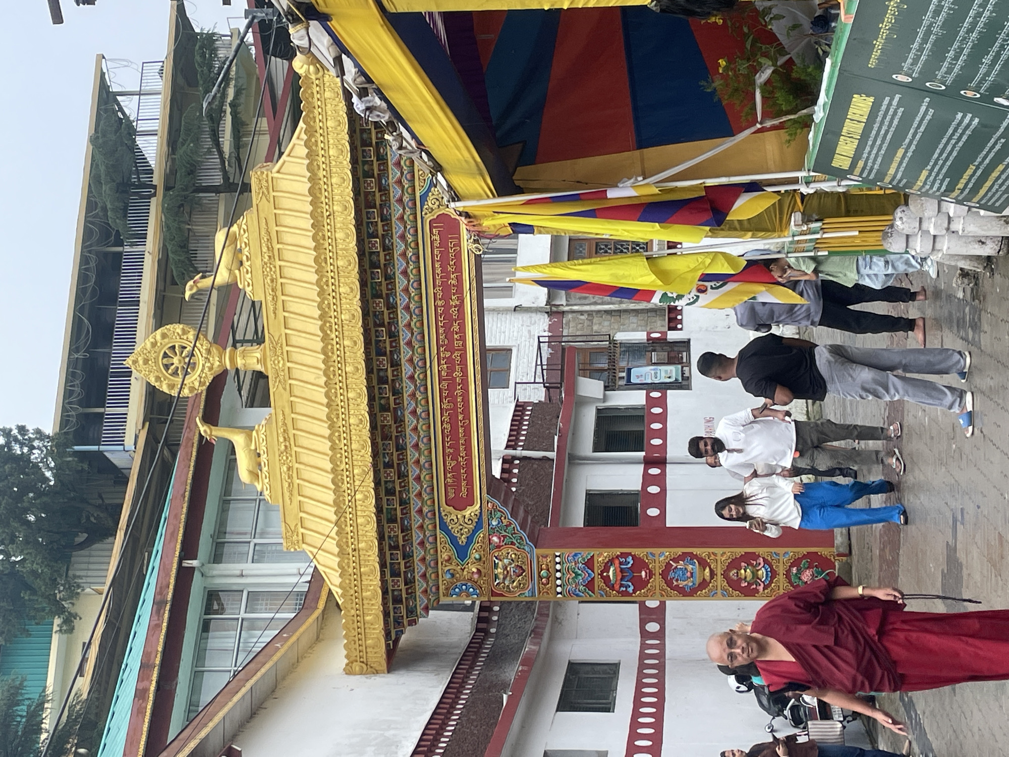 The image shows a colorful gateway at a Buddhist monastery in Dharamshala, India. The gateway is adorned with gold ornamentation, including statues of deer and a Dharma wheel. Tibetan prayer flags and banners hang from the structure, adding to the vibrant atmosphere. In front of the gateway, several people are walking. A monk in a maroon robe is in the foreground, looking towards the viewer. Other individuals, including a woman in blue pants and a white shirt, a man in a white shirt and gray pants, and a man in a black t-shirt and gray pants, are also present. The background reveals a multi-story building with a white facade and windows, typical of Tibetan architecture found in Dharamshala. The overall scene depicts a lively street in Dharamshala, likely a tourist or pilgrimage site.