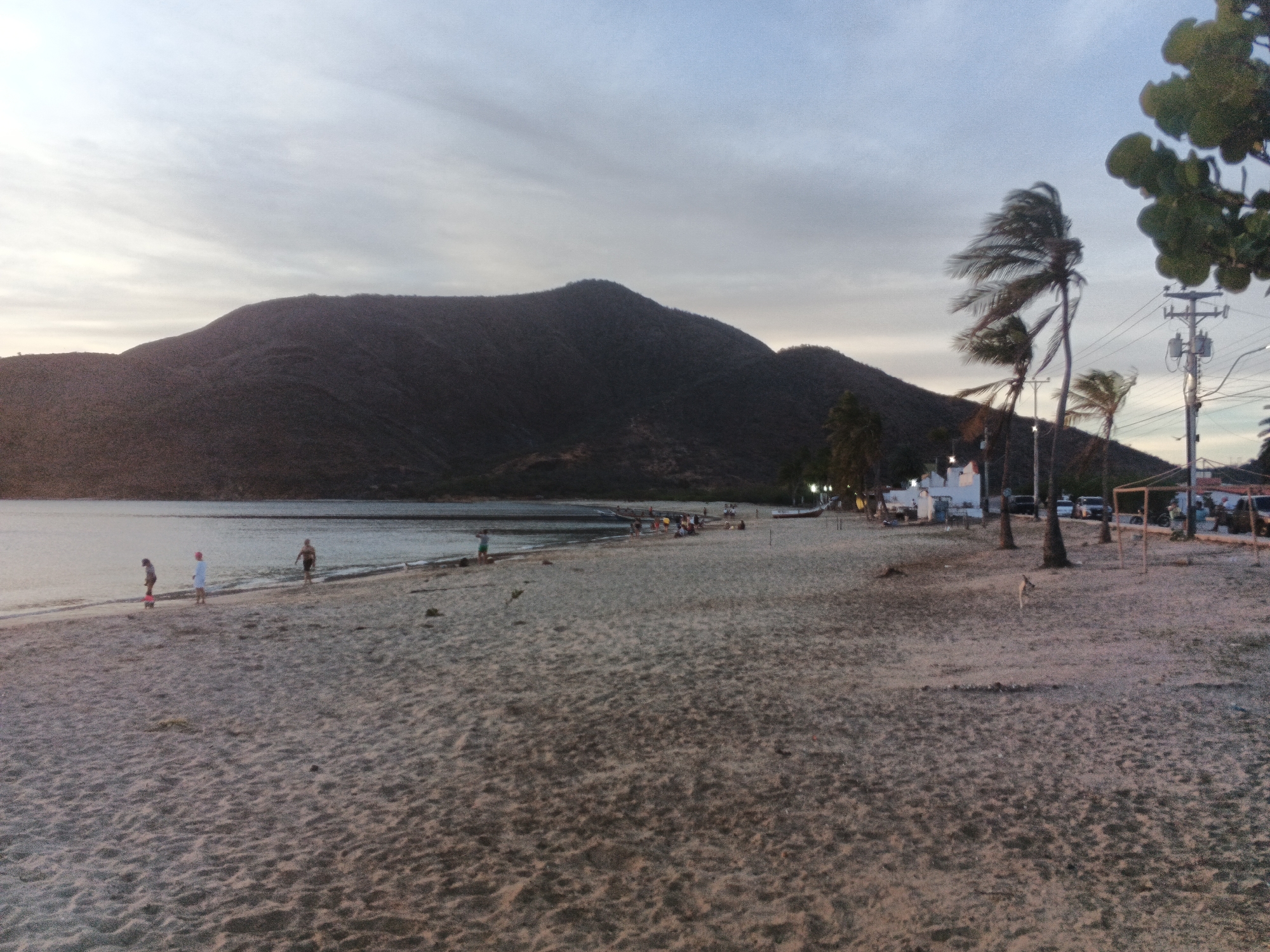 The image depicts a wide, sandy beach with several people walking near the water's edge and further back on the sand. In the foreground, the sand is textured and stretches towards the horizon. In the middle ground, a body of calm water meets the beach, and on the water's surface, there are some small boats. A dark, mountainous landscape forms the backdrop, with a slightly cloudy sky overhead. Several palm trees are visible on the right side of the image, some appearing windswept, along with a utility pole and lines. A small structure, possibly a building or shelter, is also present in the distance on the right. The lighting suggests it is either early morning or late evening.