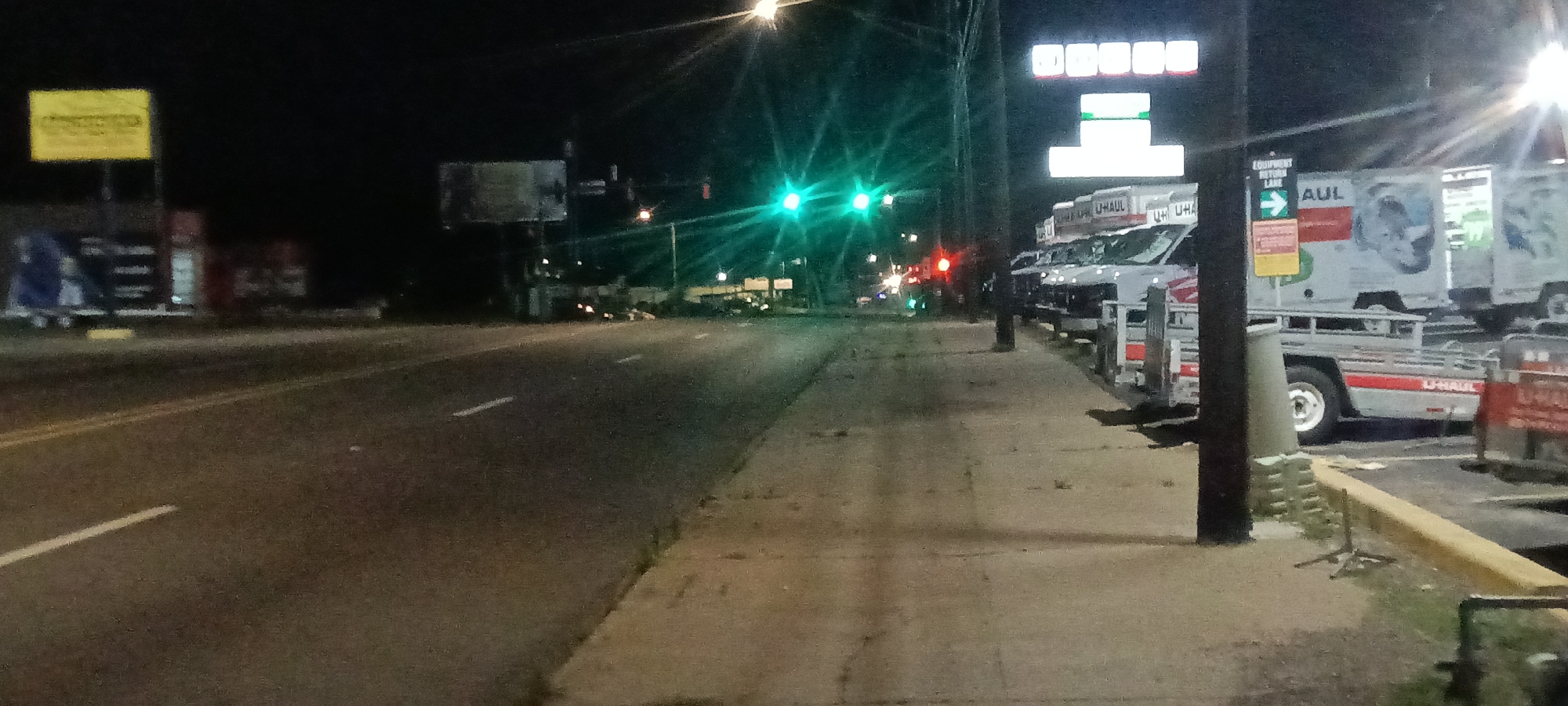 At night in Fort Smith, Arkansas, a street runs along a sidewalk with U-Haul rental trucks parked on the right. Traffic lights are green, and various signs are visible, including a yellow sign on the left. The area is illuminated by streetlights and the signs. The image is composed of a dark, clear night scene with a street and commercial trucks.