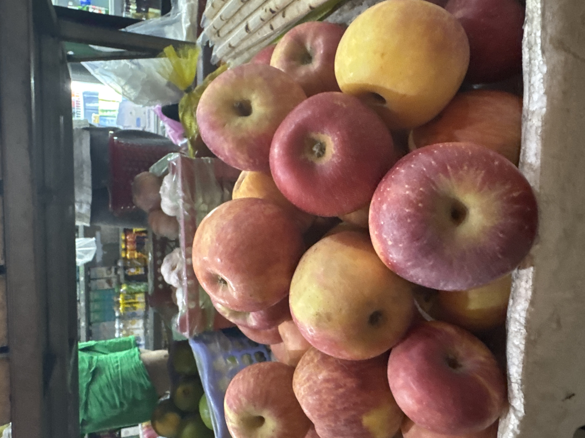 A close-up, eye-level shot focuses on a pile of red and yellow apples in a market stall in Bac Nha Trang Ward, Vietnam. The apples, some showing small brown blemishes and water droplets, are artfully arranged in a white, slightly torn, paper or cardboard container. In the background, out of focus, a person in a bright green garment is visible, along with shelves stocked with various goods, including bottles and packaged items, suggesting a bustling market environment. The lighting is somewhat dim, casting soft shadows on the fruit.