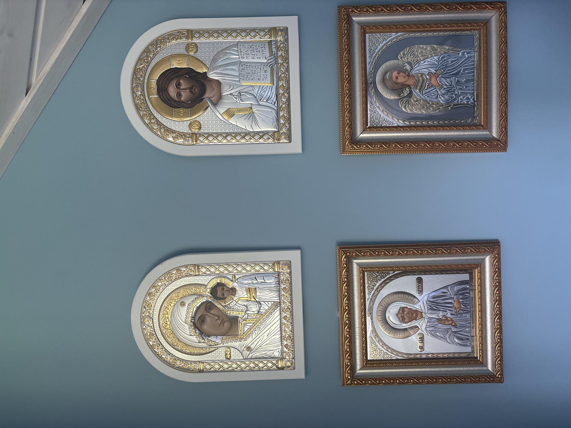 The image displays four religious icons hanging on a light blue wall. The icons are arranged in two rows, with two icons in the top row and two in the bottom row. All icons feature silver and gold detailing.
The top row icons are arched at the top. The left icon depicts the Virgin Mary holding the infant Jesus. The right icon shows Jesus Christ.
The bottom row icons are square and framed with ornate gold borders. The left icon features a woman wearing a headscarf and white robes, with her hand raised in a blessing gesture. The right icon depicts an angel with wings, also in white robes, with hands clasped over its chest.
The environment appears to be indoors, with a wooden ceiling structure visible in the upper right corner. The location is identified as Dolche Vita KP, Russia.