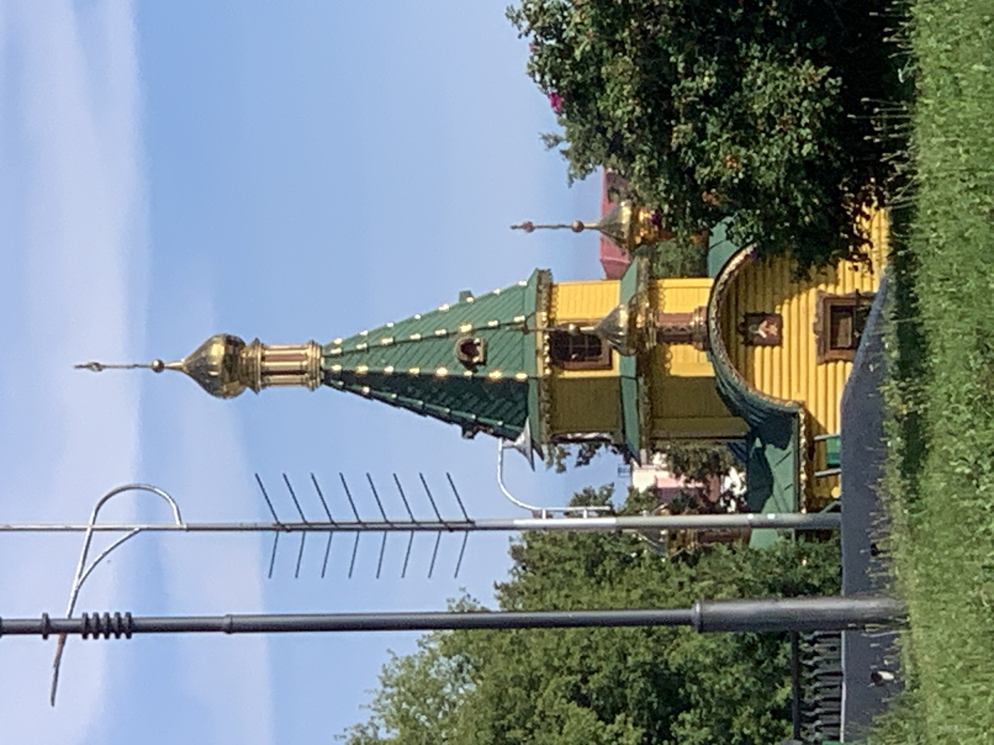 The image shows a yellow building with a green roof and golden domes, likely a church, situated among lush green trees under a clear blue sky. The building features traditional Russian architectural elements, including tiered roofs and decorative golden accents. In the foreground, tall grass and a dark fence are visible. Several utility poles and an antenna are also present on the left side of the frame. The architectural style suggests a possible location in Sluck, Belarus, a city known for its historical churches.