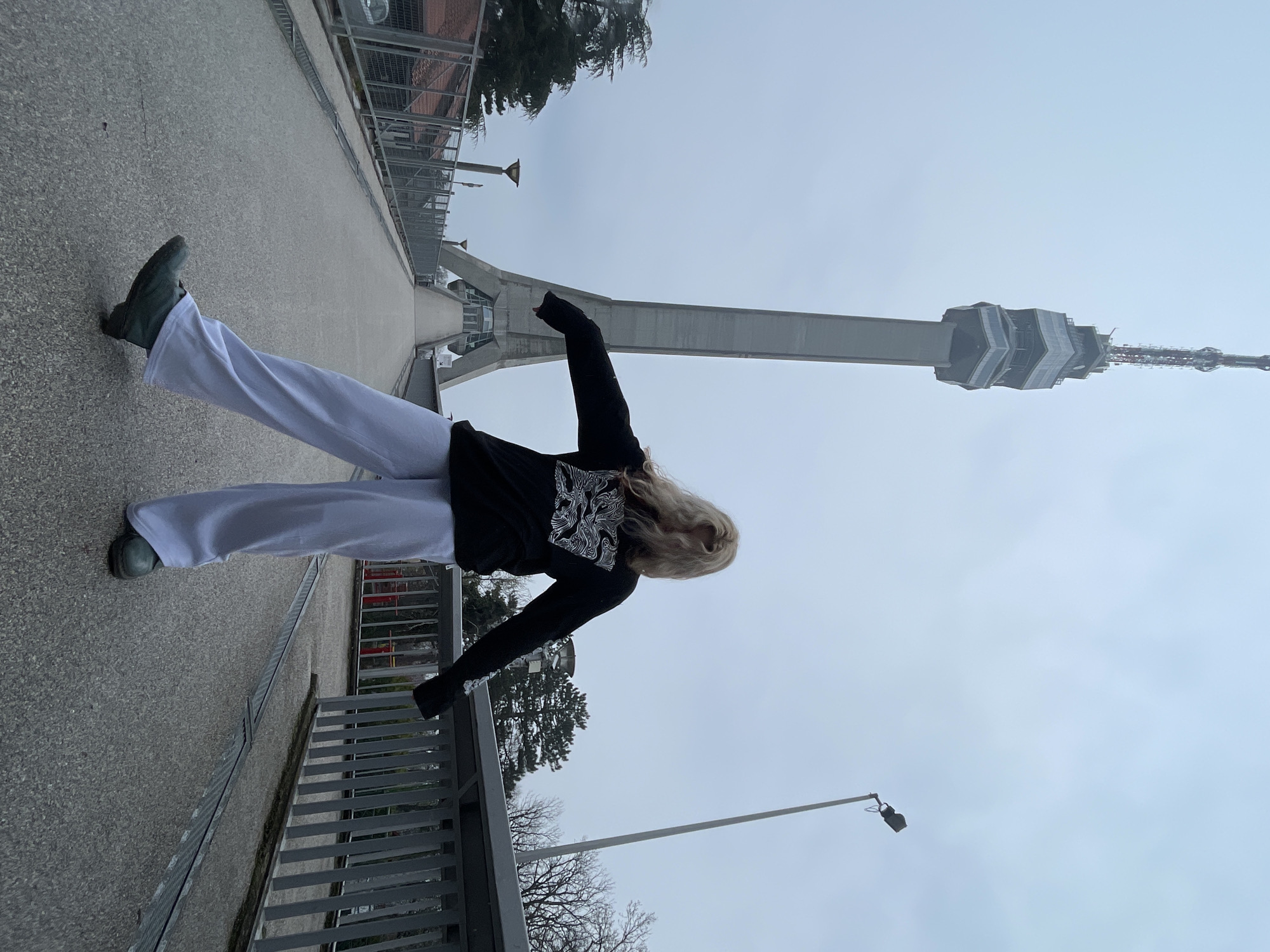 A woman in a black sweater and white pants stands on a concrete structure against the backdrop of a tall tower. The tower is the Avala Tower, located in the Vozdovac Urban Municipality, Serbia. She is facing towards the tower, with her arms outstretched. The surrounding environment is somewhat overcast.
