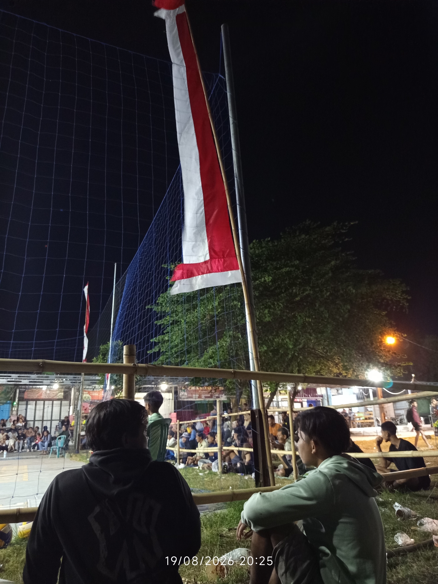 The image depicts an outdoor volleyball court at night in Canggu, Indonesia, indicated by the timestamp. The scene is illuminated by artificial lights. A red and white flag, likely the Indonesian flag, is prominently displayed on a pole, partially obscured by a volleyball net. Several people are seated and standing around the court, observing what appears to be a volleyball match or a gathering. Two individuals are in the foreground, facing away from the camera, seated on the grass. One wears a black hoodie with a graphic design, and the other wears a light green hoodie. In the background, a large tree with green foliage is visible, along with several stalls or buildings, some with illuminated signs. The overall atmosphere suggests a community event or local sporting activity taking place after dark.
