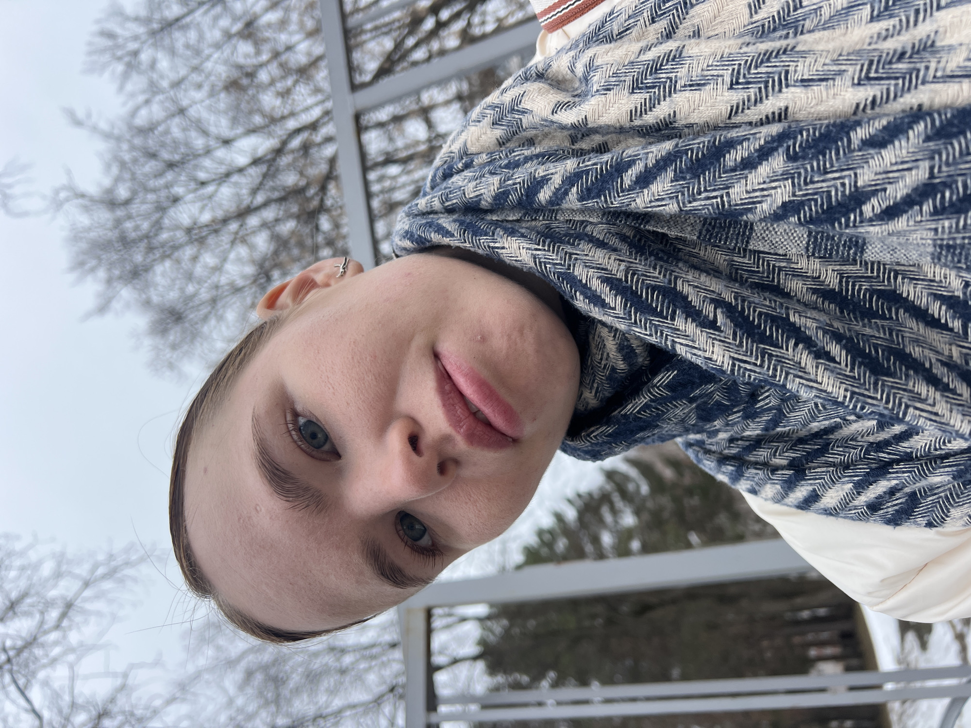 A young woman is outdoors, wearing a blue and white herringbone scarf. The location is suggested to be Chaikovsky, Russia, and the background includes trees and a metal railing. The sky is overcast. The woman is looking directly at the camera.