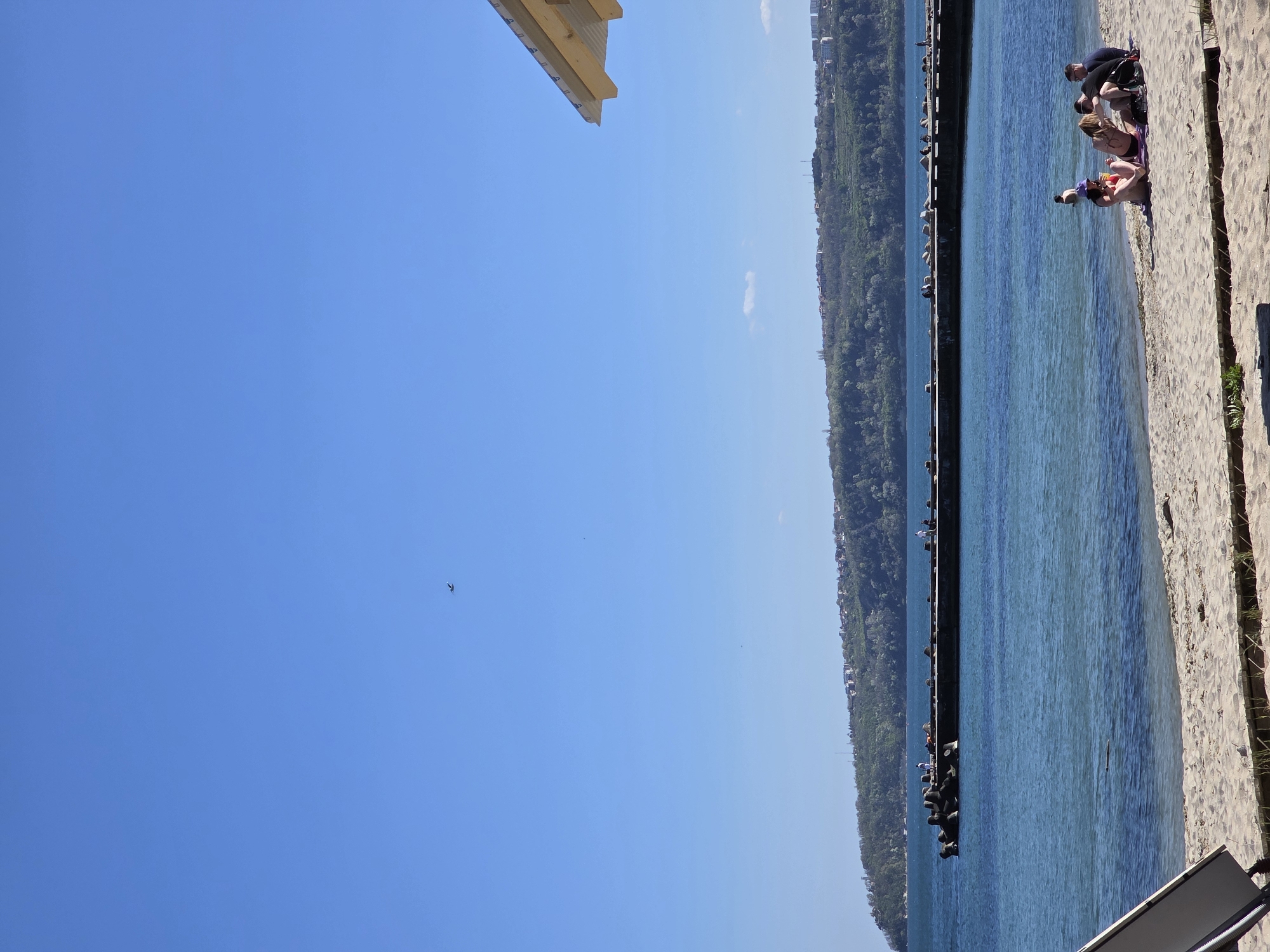 The image shows a view of the beach and sea in Varna, Bulgaria. The camera angle is oriented sideways. In the right portion of the frame, several people are seated or lying on the sand near the water's edge. Adjacent to the beach, a long pier extends into the sea, with trees on the opposite side. The sky is clear and blue. A partial view of a wooden structure is visible in the upper-left corner.