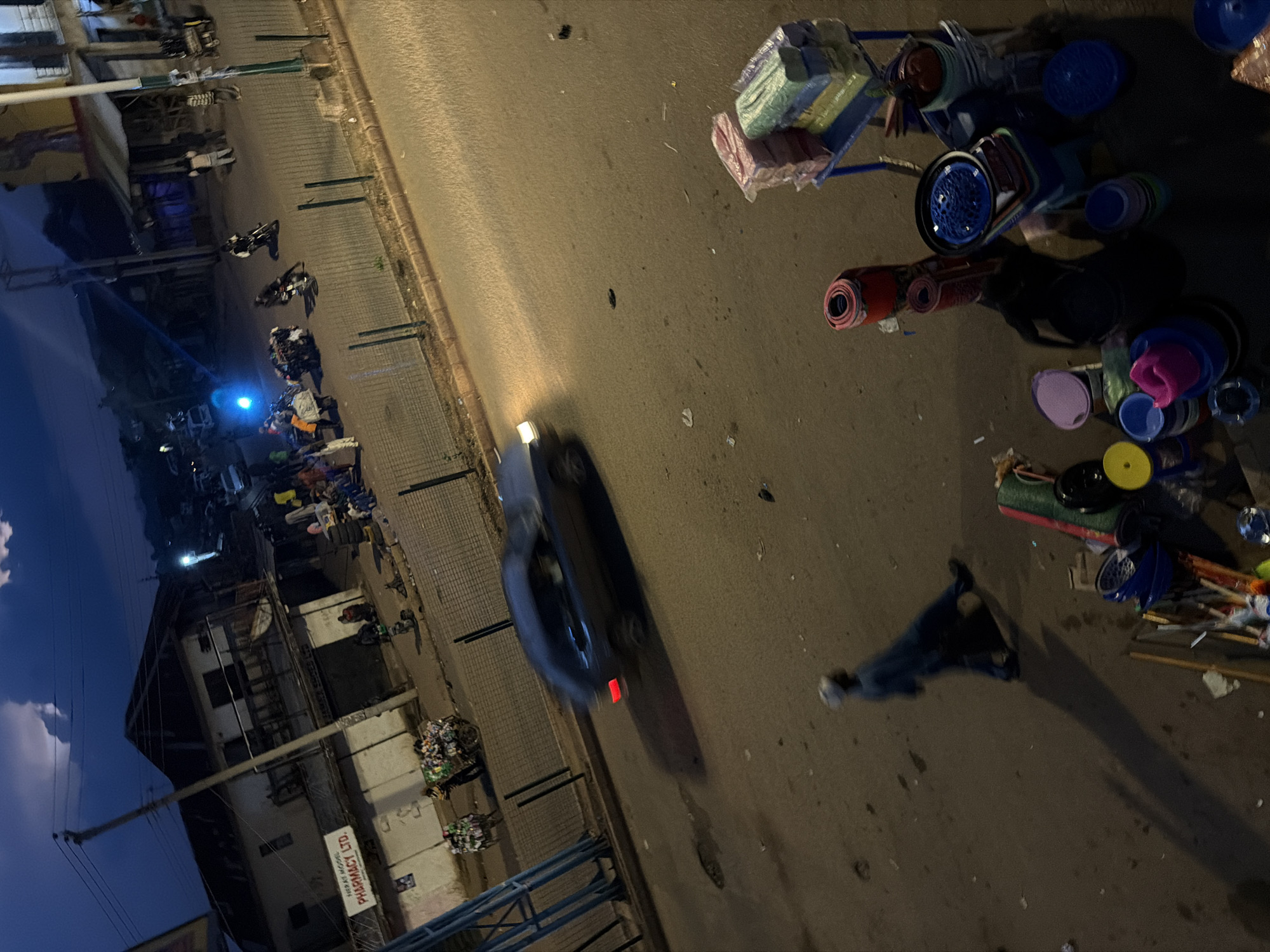 The image depicts a street scene at dusk or night in Bukuru, Nigeria. The perspective is from above, looking down at an angle. In the foreground, various objects are scattered on the asphalt, including rolled-up mats, baskets, plastic containers of different colors (blue, yellow, pink, purple), and what appears to be a cart laden with packaged goods. A person, wearing dark clothing and a blue garment draped over their shoulders, is visible, casting a long shadow on the ground.
Further into the scene, a dark-colored car is in motion, with its headlights and taillight illuminated, suggesting it is driving on the road. The road surface is a mix of asphalt and possibly paved stones near the buildings. To the left, buildings line the street. One building has a sign with visible text that reads "NEXT LEVEL PHARMACY LTD." The buildings appear to be of multiple stories with balconies and windows.
Several motorcycles are parked or in motion near the buildings. Some people are visible around the motorcycles and in front of the businesses. The overall impression is of a busy street, possibly a market or commercial area, in the evening. The lighting suggests artificial illumination from streetlights and the vehicles, combined with the fading natural light. The sky on the far left shows clouds and a deep blue hue, indicative of twilight. The presence of trash or debris is noticeable in some areas, particularly near the buildings.
