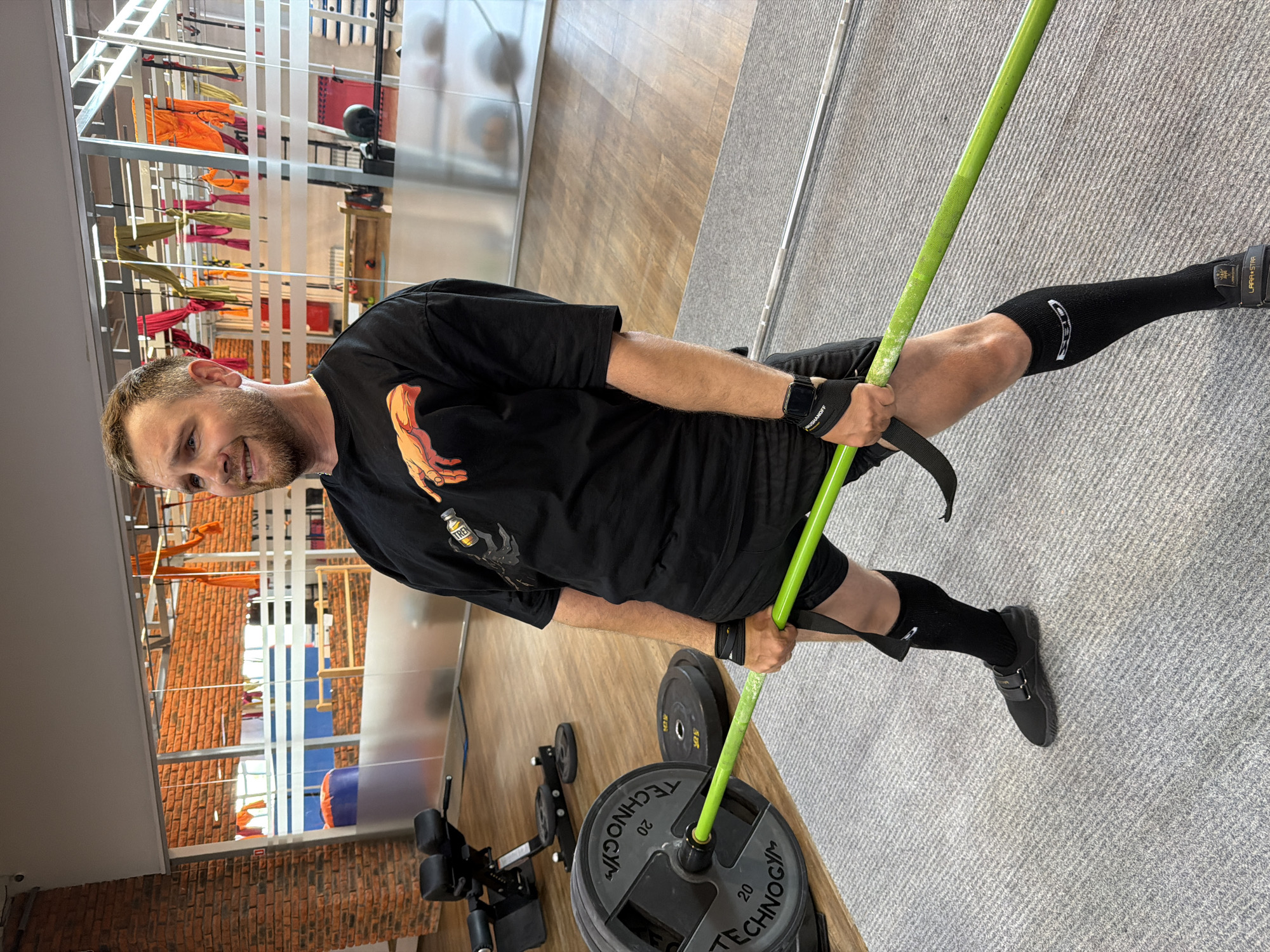 A man is performing a deadlift in a gym. He is wearing a black t-shirt with an orange design on the front, black shorts, black socks, and black athletic shoes. He is holding a green barbell with both hands. The gym has a brick wall on the left, and mirrors with acrobatic equipment hanging behind them on the right. The floor is covered in a grey carpet. The scene appears to be in the city of Chaikovsky, Russia, as indicated by the presence of a Technogym brand barbell, a common fixture in fitness facilities.