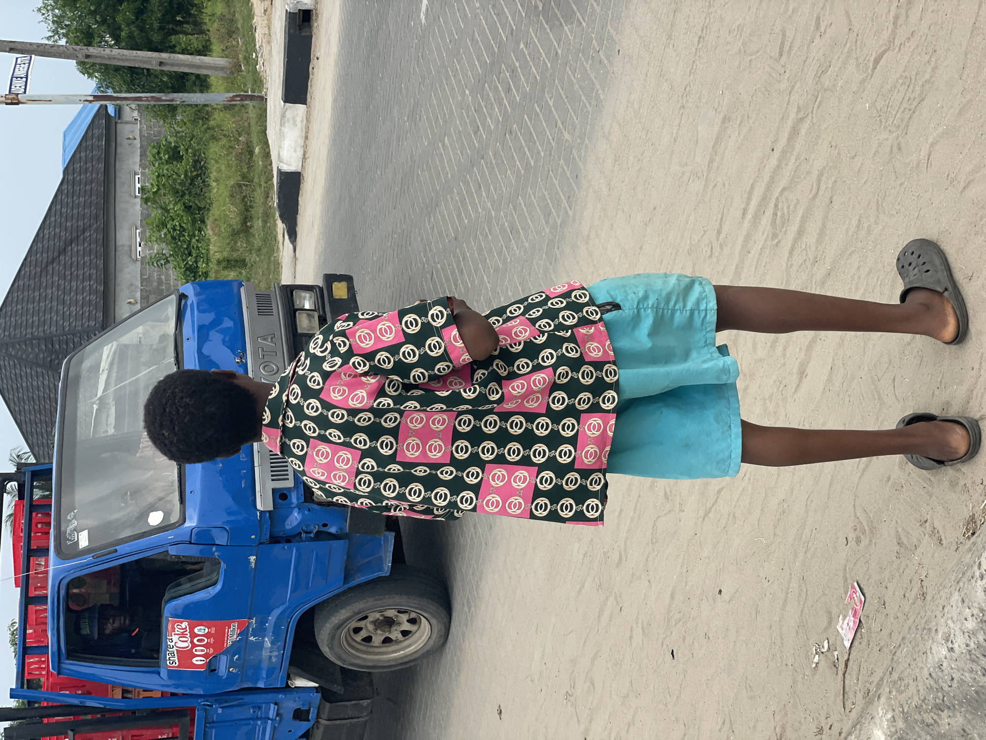 A medium shot depicts a young male standing next to a blue Toyota truck in Badagry, Nigeria. The truck is parked on a sandy shoulder adjacent to a paved road marked with a black and white curb. The truck's flatbed is stacked with red crates. The young man is wearing a patterned shirt with pink and white designs on a dark green background and light blue shorts. He has dark skin and short, curly hair. Gray sandals adorn his feet. To the left, a building with a gray roof and exterior walls is visible, alongside a street sign.