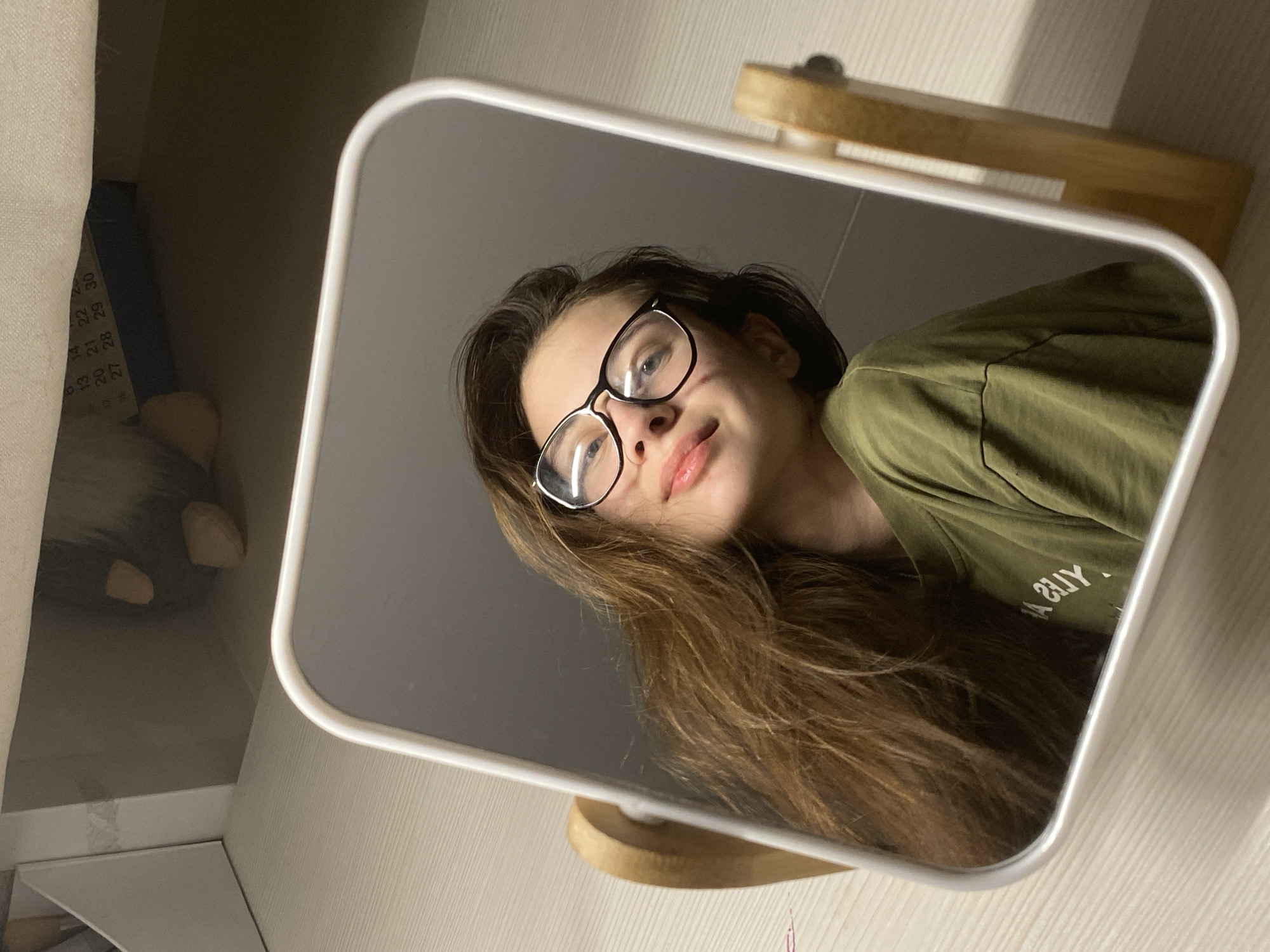 A young person, likely female, is captured in a mirror selfie in an indoor setting in Votkinsk, Russia. The image is taken from a low angle, looking up into a rectangular mirror with rounded white edges, which is placed on a wooden stand. The subject, positioned centrally within the mirror's reflection, has fair skin, long brown hair, and is wearing black-framed glasses. She is looking upwards into the camera with a gentle smile and slightly parted lips.
She is wearing an olive-green t-shirt, which has mirrored white text visible on the chest area. The text appears as "S?UY. A" and, when unmirrored, could spell "A. ZUY" or something similar.
In the background of the mirror's reflection, several details are visible:
* To the left, partially obscured by a light-colored fabric or curtain, is a calendar or schedule with numbers visible, including "13, 14, 20, 21, 22, 27, 28, 29, 30". Below this, a dark object and a partial view of a plush toy resembling a gnome with fuzzy texture and light-colored 'feet' or 'hands' are seen.
* To the right, next to the mirror, there are light-colored surfaces, possibly a wall or shelf, and some small, light-colored, irregularly shaped objects, possibly decorative stones.
The lighting appears soft, possibly from a natural source with some ambient room light, creating subtle shadows. The overall scene suggests a relaxed moment, with the subject taking a casual mirror photograph.
