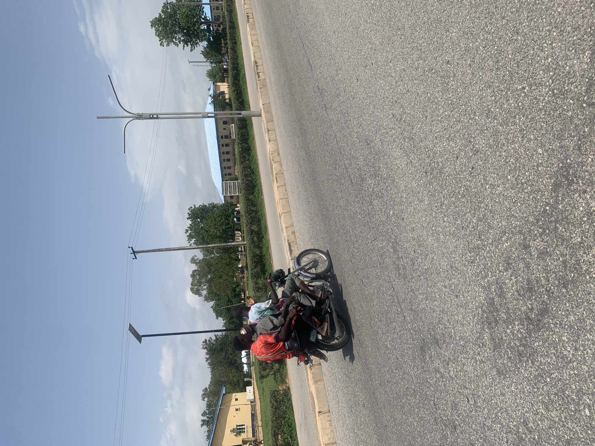 In Jalingo, Nigeria, three young men are riding on a motorcycle on a paved road. The rider in the front wears a red shirt, the middle passenger is in a grey shirt and hat, and the person in the back is wearing a tie-dye shirt. The motorcycle has a license plate reading "KM 0509". In the background, there are buildings with blue roofs, trees, streetlights, and utility poles. The sky is partly cloudy.