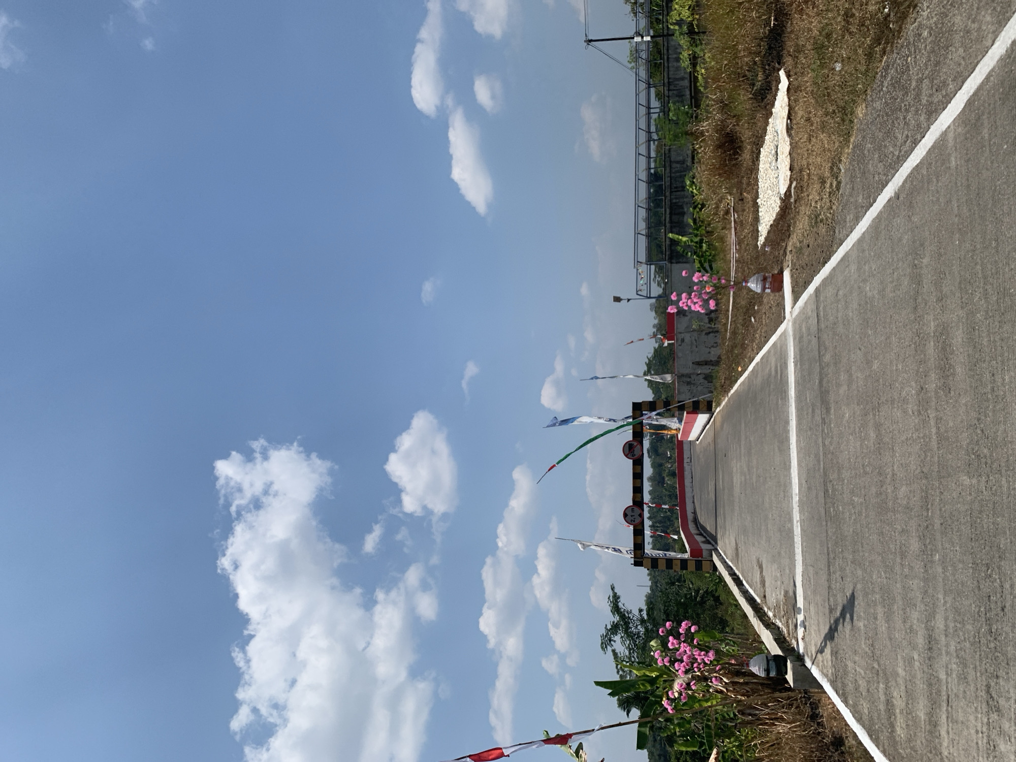 This image depicts a road leading towards an archway in Ciamis, Indonesia. The road is paved with concrete and has white markings. The archway is decorated with black and yellow stripes, and flags are displayed on either side. There are also two circular signs on the archway, one indicating a height restriction of 2.1 meters and the other a warning for trucks. Pink flowers in a plastic bottle are placed on the side of the road, near the entrance. In the background, there are trees and a fence. The sky is blue with scattered white clouds, suggesting a clear day. The scene appears to be a public thoroughfare, possibly a gateway to a specific area or attraction.
