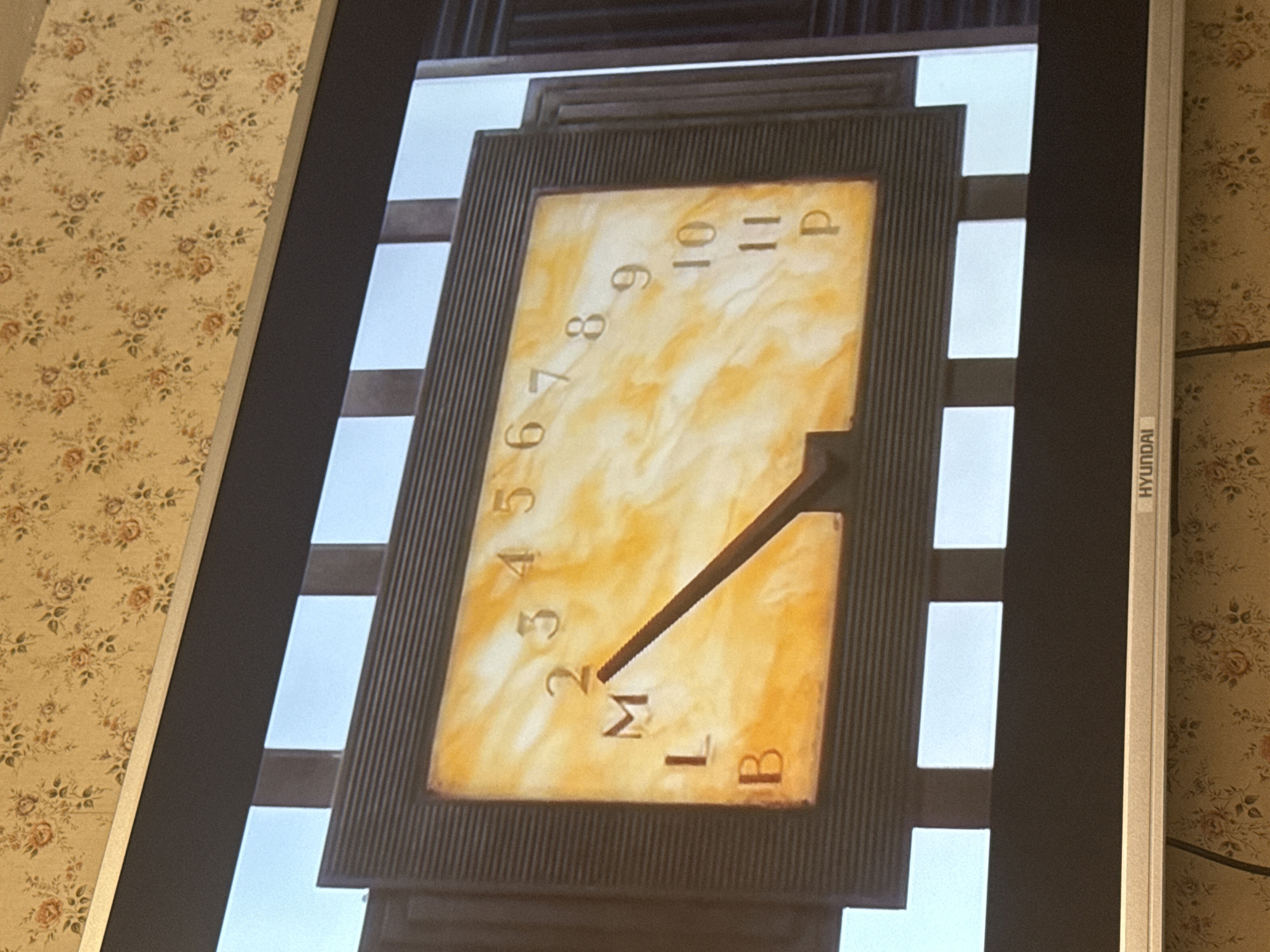 The image shows a photograph of a screen displaying a sundial clock. The clock face is rectangular and framed, with numbers ranging from 2 to 11, and letters "M," "L," "B," and "P." A gnomon casts a shadow indicating the time. The screen itself is a Hyundai brand monitor. The monitor is situated in a room with floral wallpaper. The location of the scene is in Mytishchi, Russia.