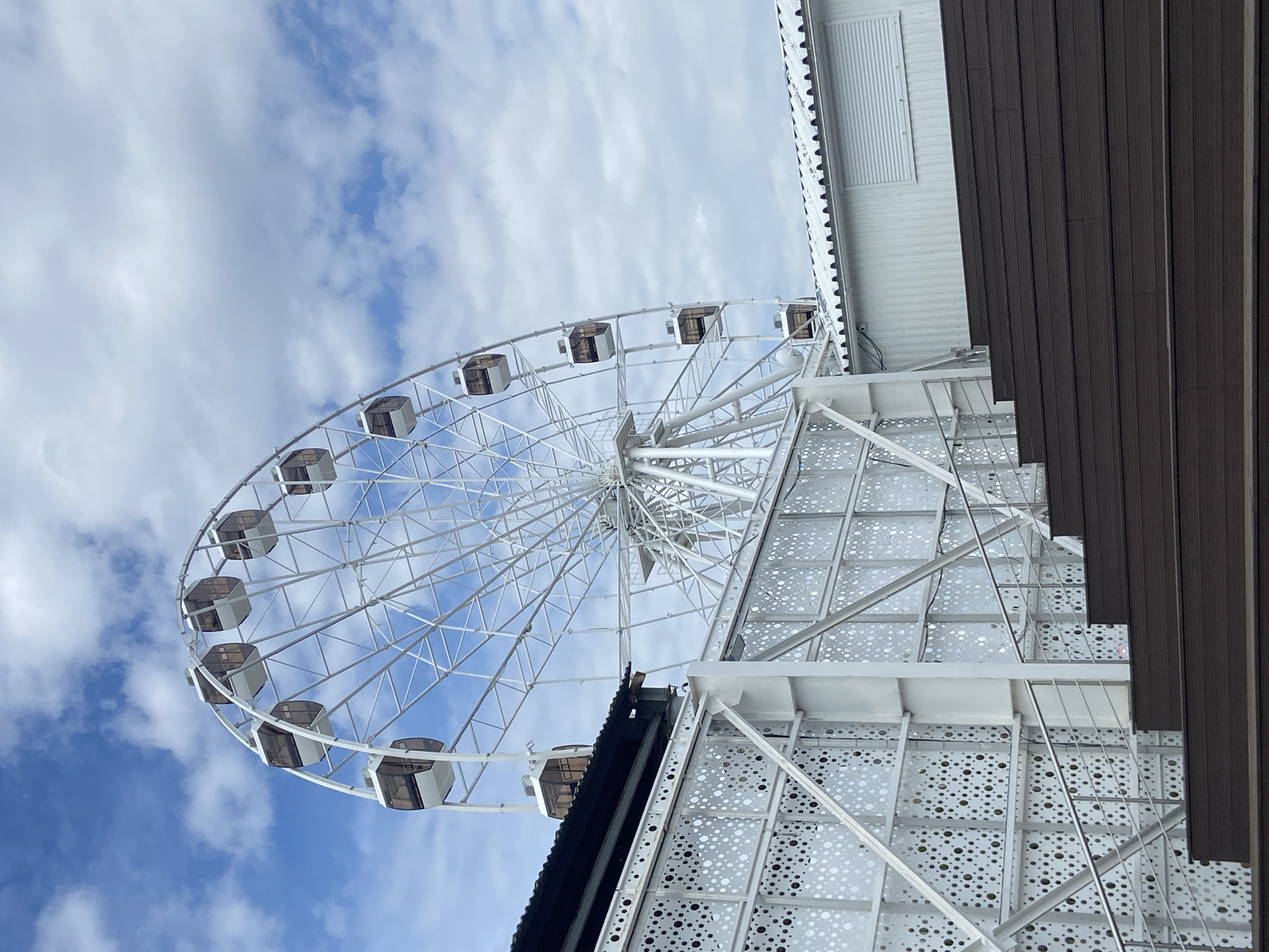 The image shows a Ferris wheel against a cloudy sky. The Ferris wheel is white, with enclosed gondolas. Adjacent to the Ferris wheel is a building structure, also white with a patterned facade, accompanied by brown wood slats. The perspective is looking up at the Ferris wheel from ground level. It appears to be in Saratov, Russia.