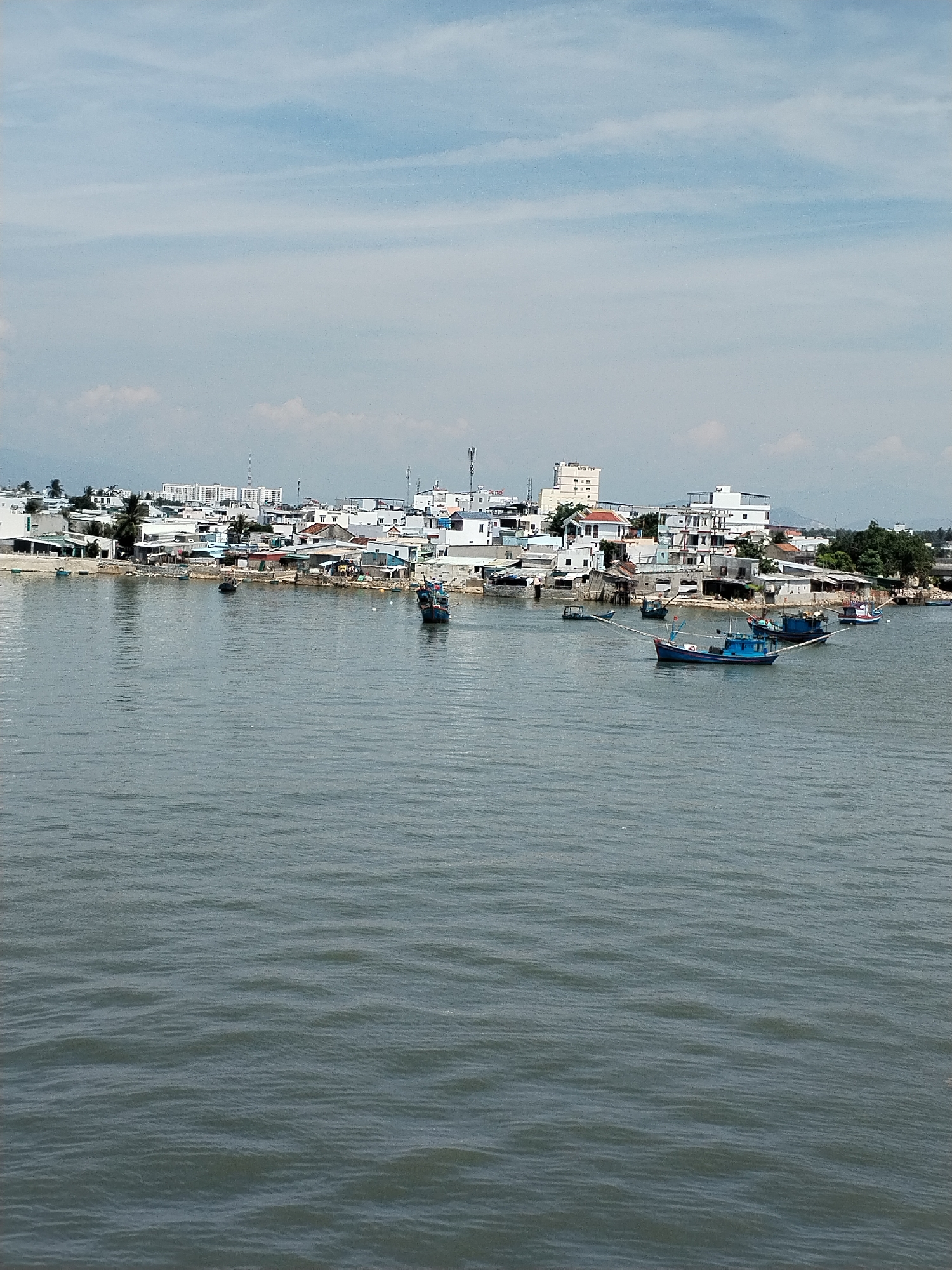 The image depicts a body of water in front of a coastal city skyline. The water is choppy with small waves. Several boats are present on the water, including at least three blue fishing boats in the foreground. In the background, a city with a mix of low-rise buildings and taller structures is visible along the coastline. The sky is overcast with wispy clouds. The scene is likely from the city of Nha Trang, Vietnam, a popular coastal tourist destination known for its beaches and fishing industry.