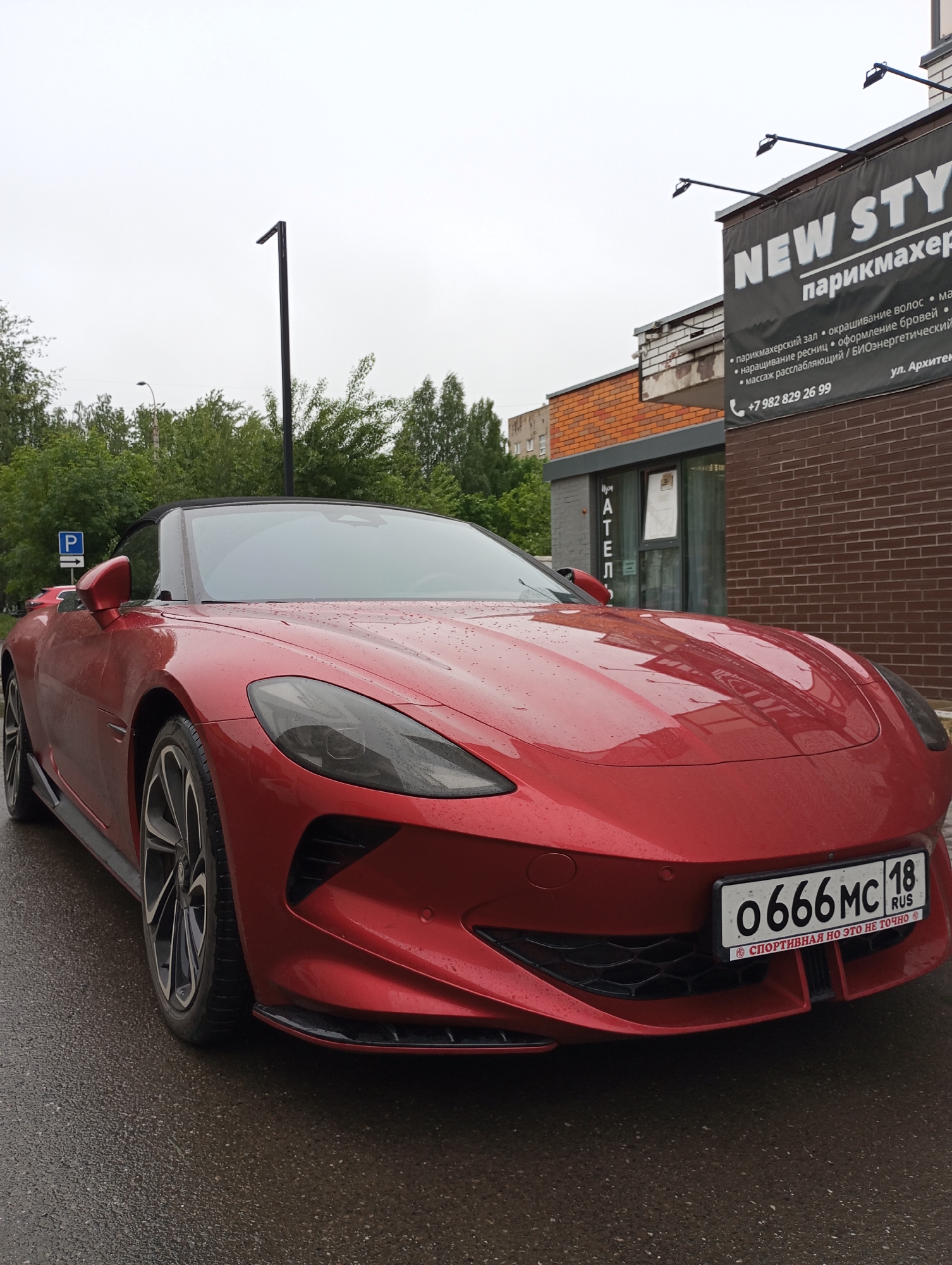 The image shows a red sports car parked on a wet street in Izhevsk, Russia. The license plate number is O666MC 18. Behind the car is a building with a sign advertising "NEW STYLE парикмахер," a hair salon. The sign includes a phone number and address (ул. Архитек). A parking sign is visible to the left of the car. The weather appears overcast and rainy.