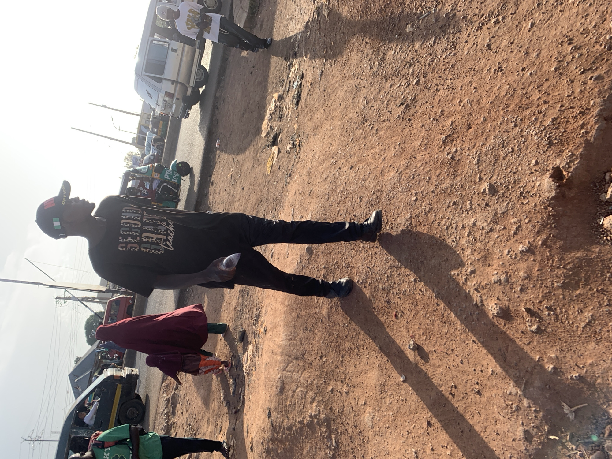 A street scene in Gura Top, Nigeria. A man with a black t-shirt and a hat is walking on a dirt shoulder beside a road. Other people walk along the road, some in hijabs. Vehicles, including cars and tuk-tuks, are parked and driving along the road. Power lines are visible. Trash is visible in the dirt shoulder.